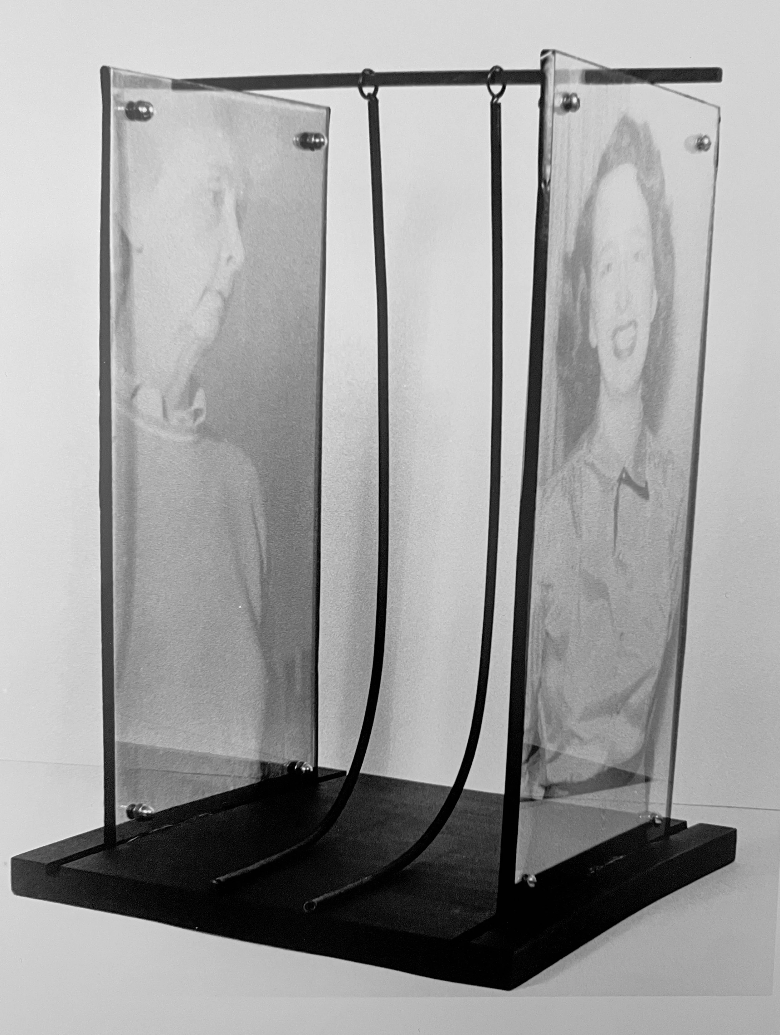 A modern art sculpture with two female portraits on glass panels, mounted on a black base with metal frame, featuring three black cords hanging from horizontal bar by artist, Karen Serago.
