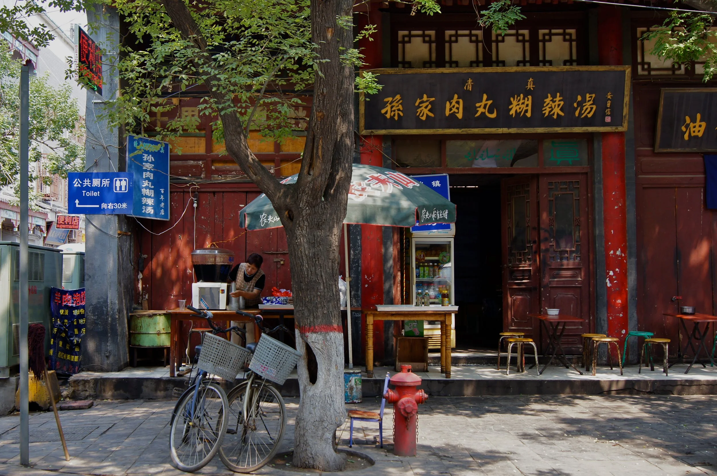 Street scene with a tree in foreground, outdoor food stand with a woman preparing food, two bicycles parked in front, and traditional Asian signage on building in the background, photographed by Karen Serago.