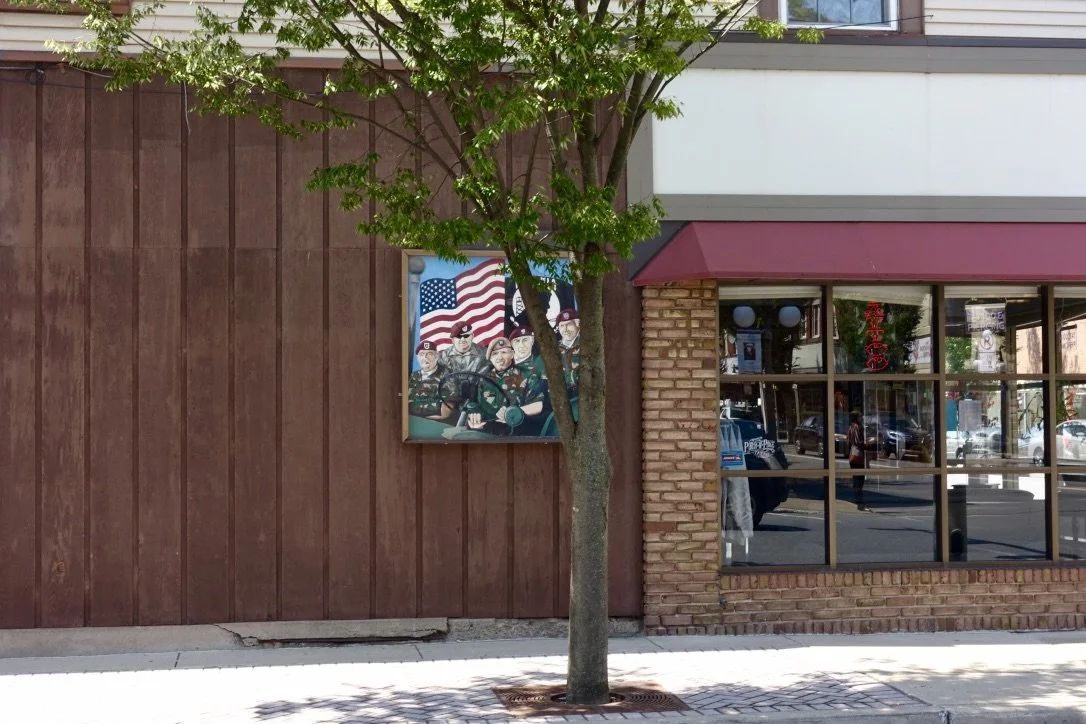 A mural of four soldiers in military uniforms with an American flag in the background, mounted on a brown wooden wall outside a building, next to a window with red awning and a tree providing shade, photographed by artist, Karen Serago.