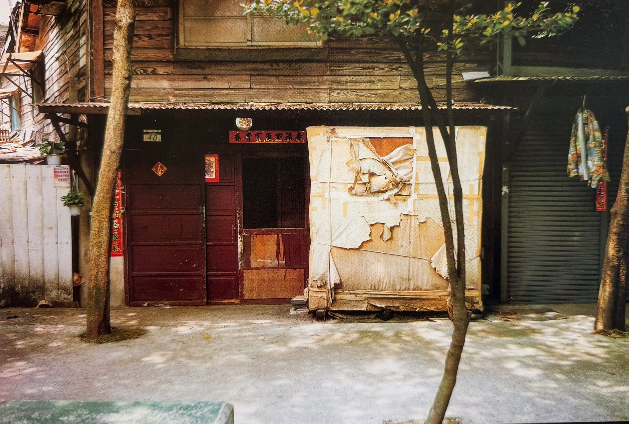 An old, worn building with a corrugated metal roof, surrounded by trees and a dirt ground. The front features a red wooden door, a tiled banner with Chinese characters, and peeling paper or wallpaper on part of the wall. There is a small potted plant on a white fencing and various signs or decorations. The overall scene appears aged and somewhat neglected.