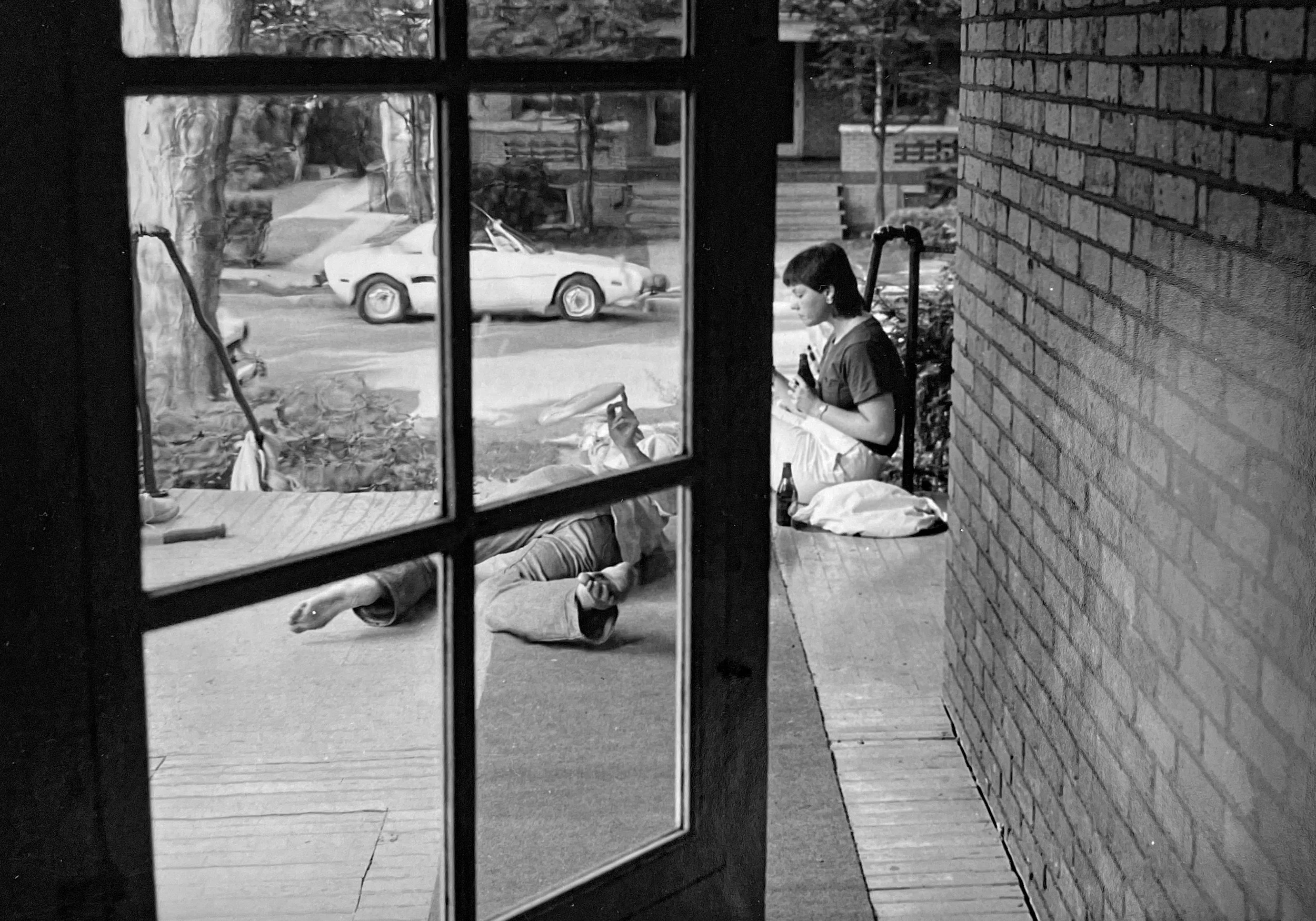 View through a window showing a person laying on the ground with a woman beside her. In the background, there is a car and a street scene with houses, photographed by visual artist, Karen Serago.