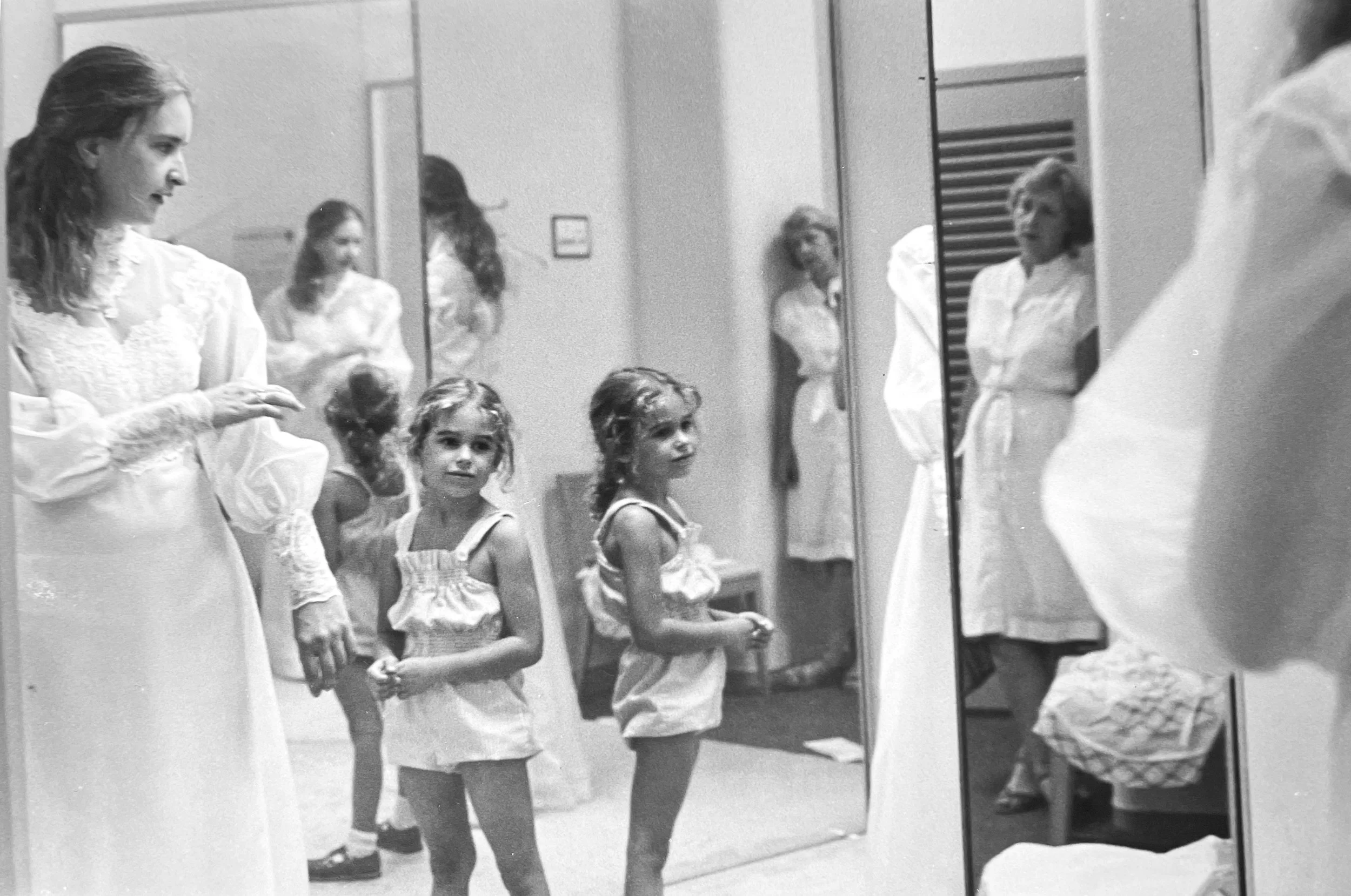 A woman and two young girls standing in front of a mirror, getting ready or preparing, with a woman in the background also visible, photographed by visual artist, Karen Serago.