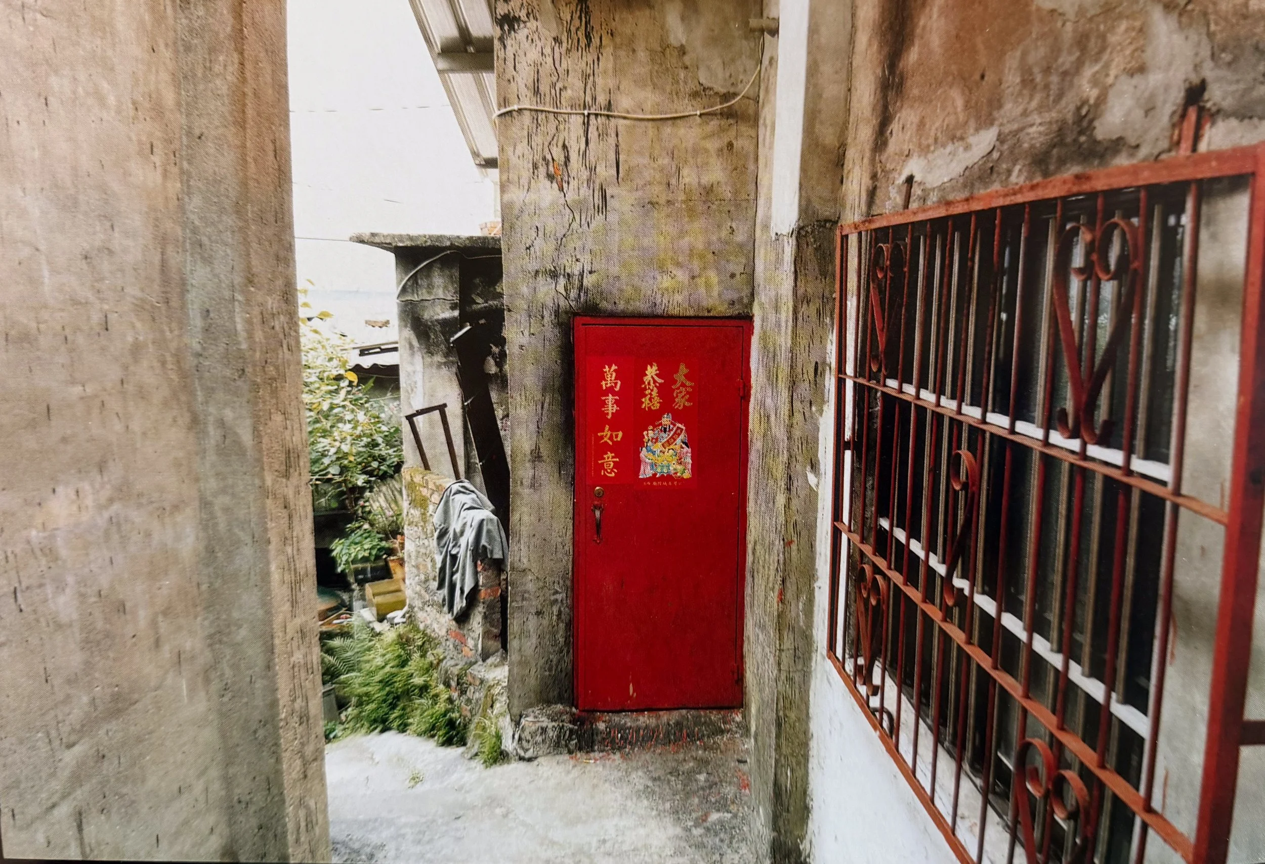 An alleyway with a red door featuring Chinese characters and a cartoon image, enclosed by concrete walls and a barred window, with some greenery and personal items visible outside, photographed by visual artist, Karen Serago.