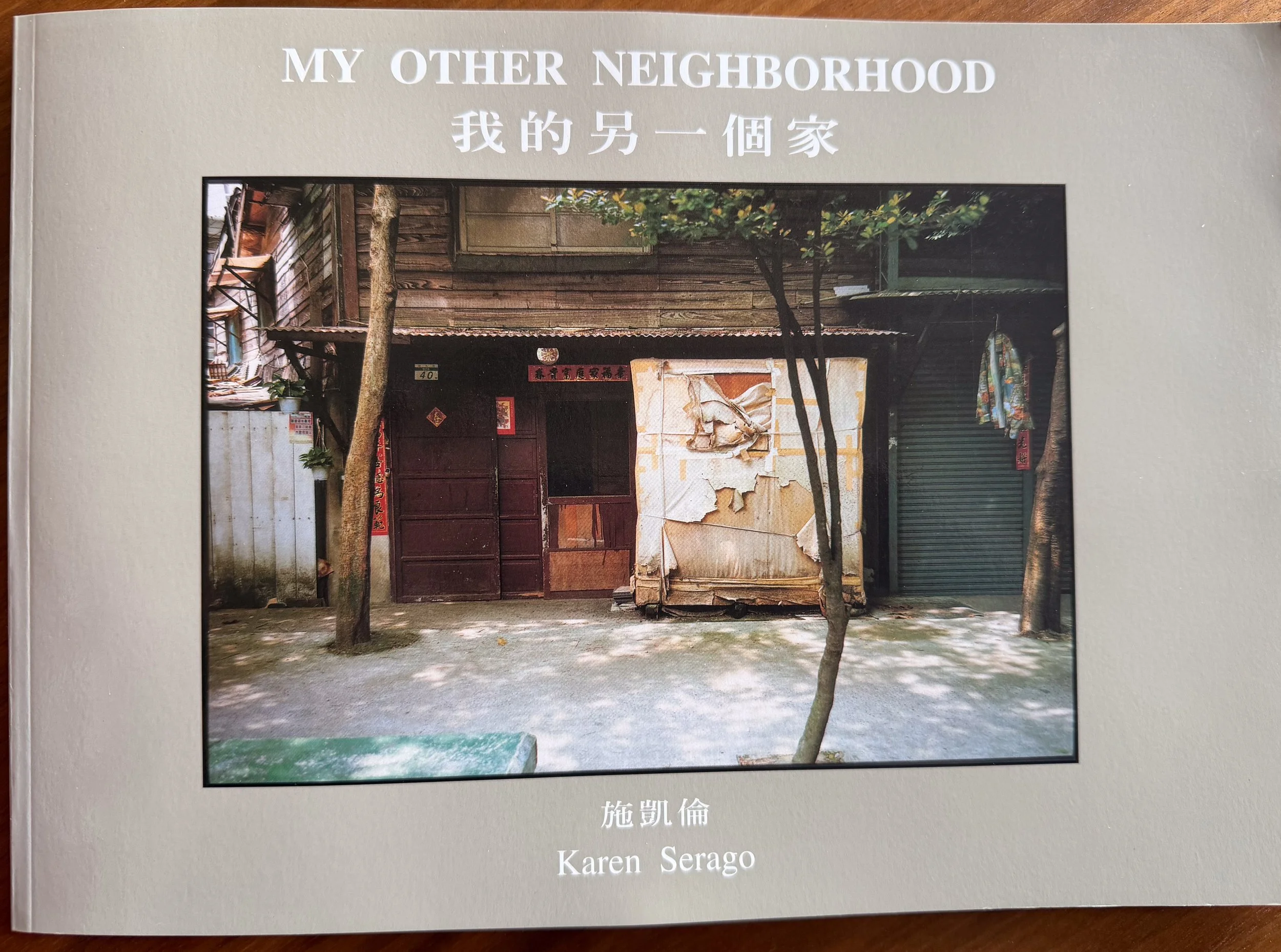 A book cover titled 'My Other Neighborhood' by Karen Serago, visual artist and author, featuring a photograph of an old, weathered building with peeling paint, a red door, and a small tree outside.