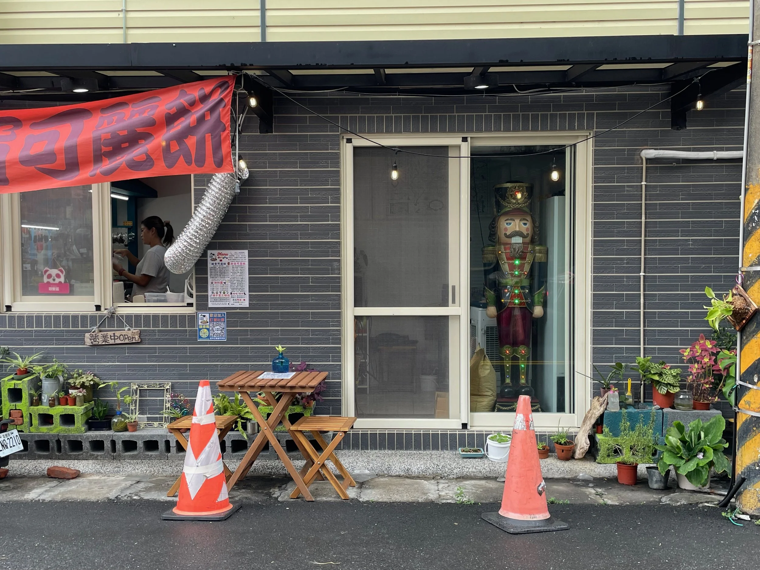 A storefront with a red banner and a large toy soldier figure inside, seen through the glass door. The exterior has potted plants, small wooden tables, orange traffic cones, and a sign indicating it is open, photographed by artist, Karen Serago.