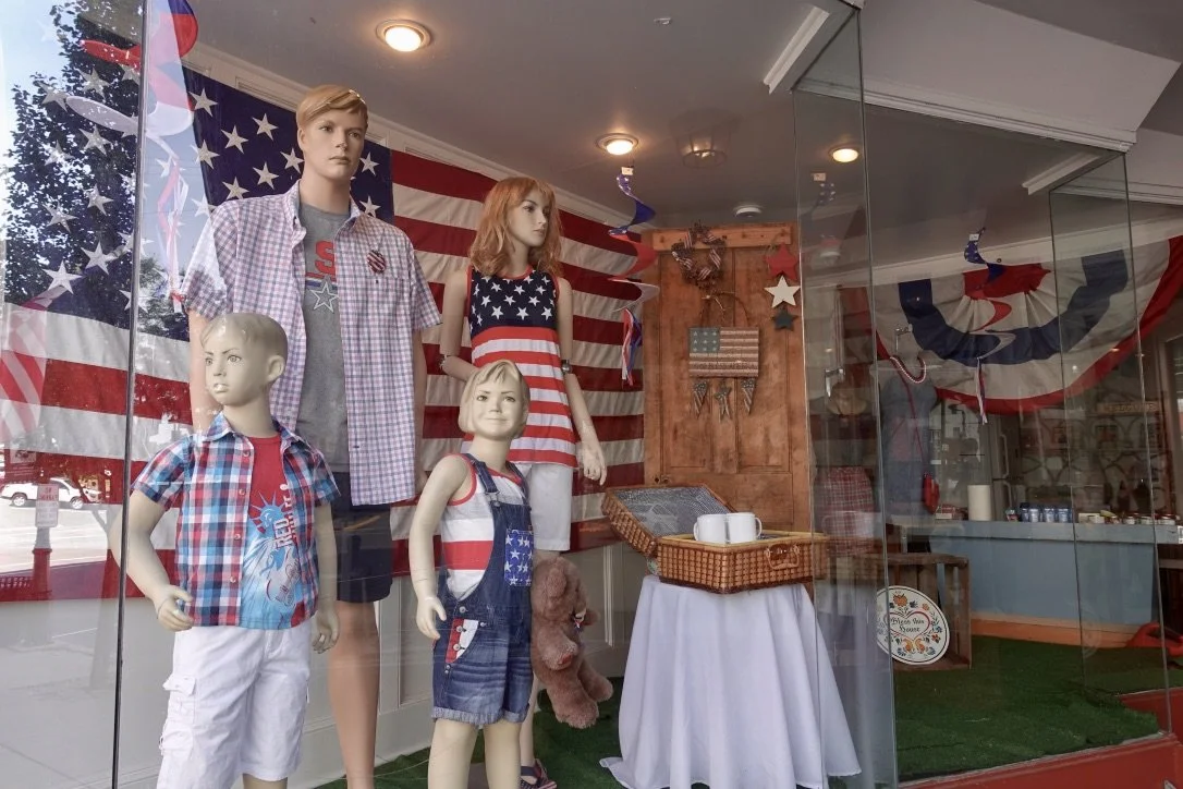 Display window with mannequins dressed in patriotic American-themed clothing, red, white, and blue decorations, and an American flag background, celebrating American pride photographed by artist, Karen Serago.