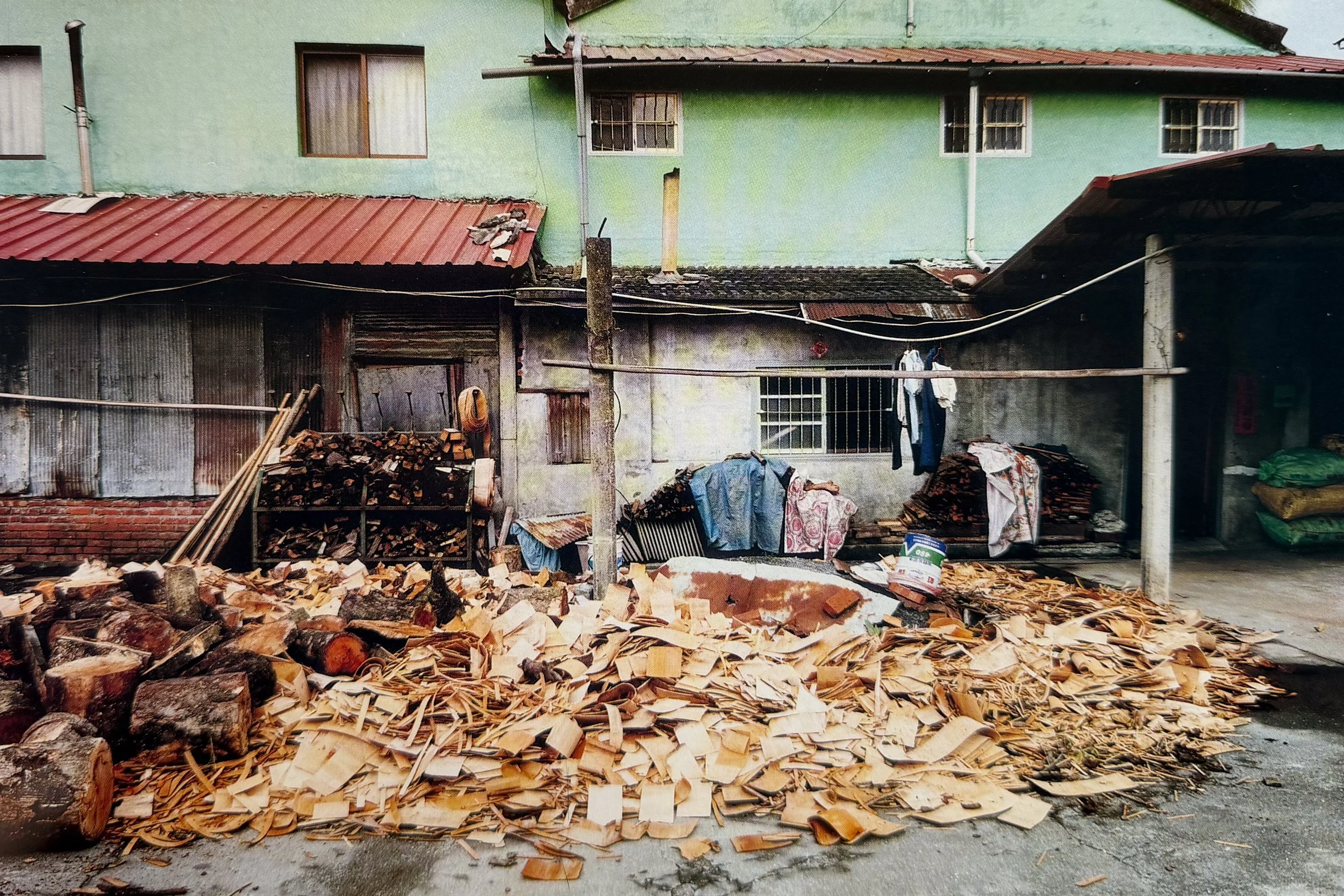 A pile of chopped firewood on the ground in front of a worn house with a green upper wall and corrugated metal lower walls. Clothes are hanging on a line near the house. Photographed by visual artist, Karen Serago.
