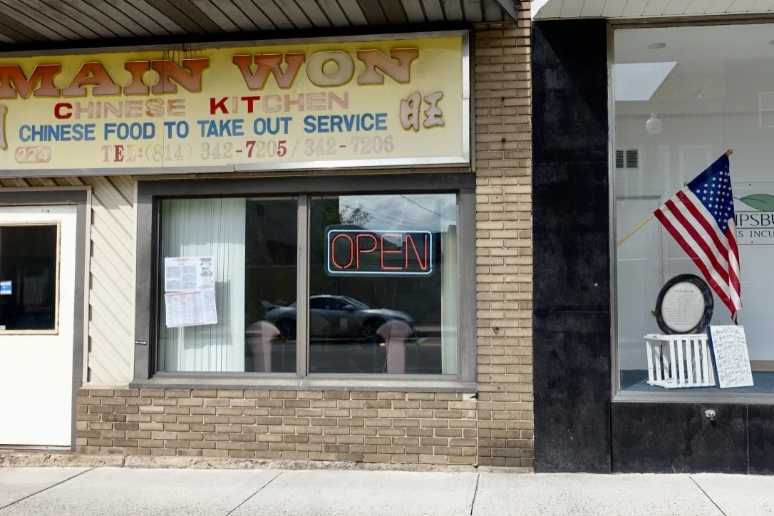 Face of a Chinese restaurant called 'Main Won Chinese Kitchen,' with a neon 'Open' sign in the window, and a display with an American flag beside it, photographed by artist, Karen Serago.