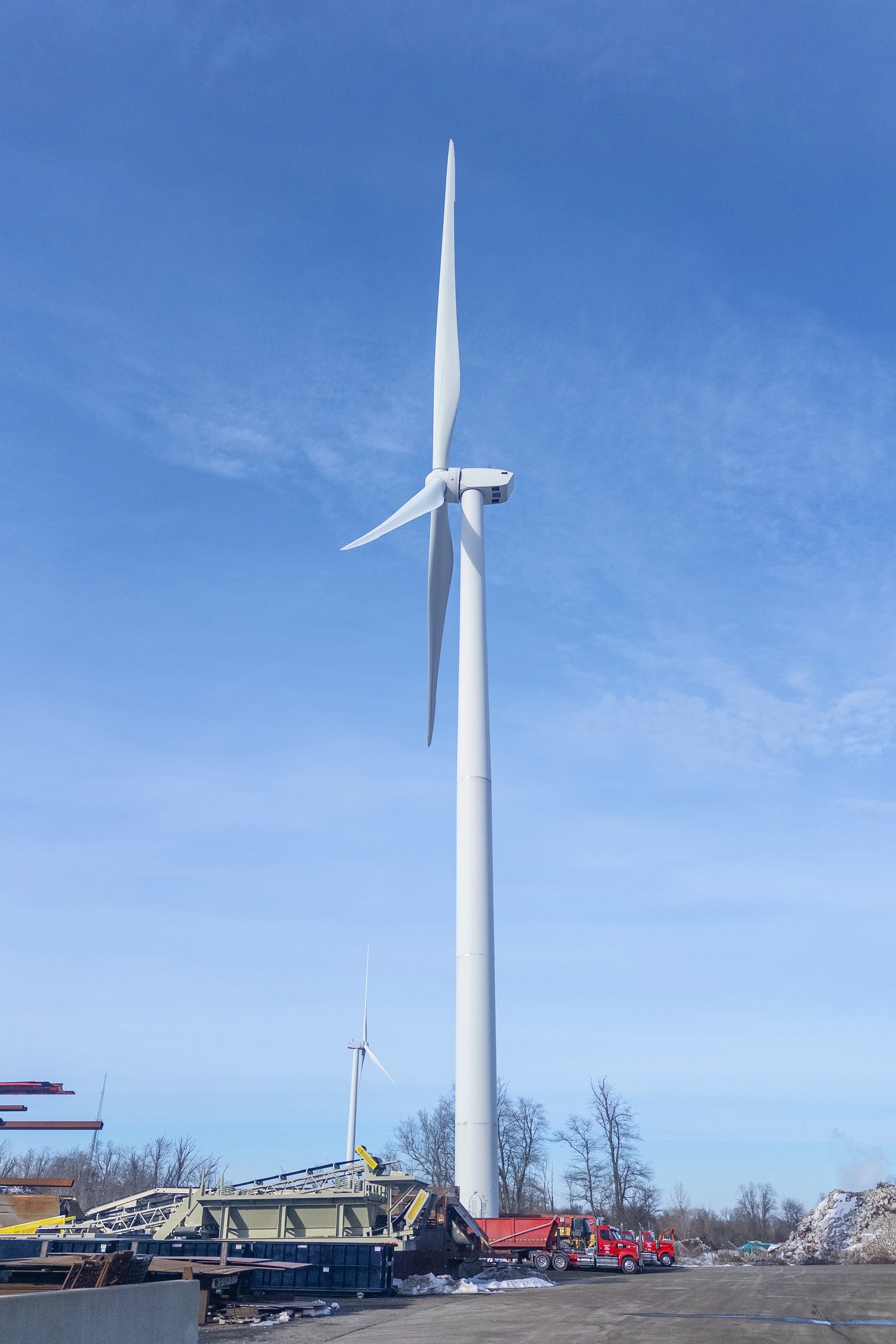 Image depicts that grand scale of a wind turbine in Johnston, RI against the clear blue sky, compared to a large 18-wheel red truck along it's base. Taken on 2.15.26