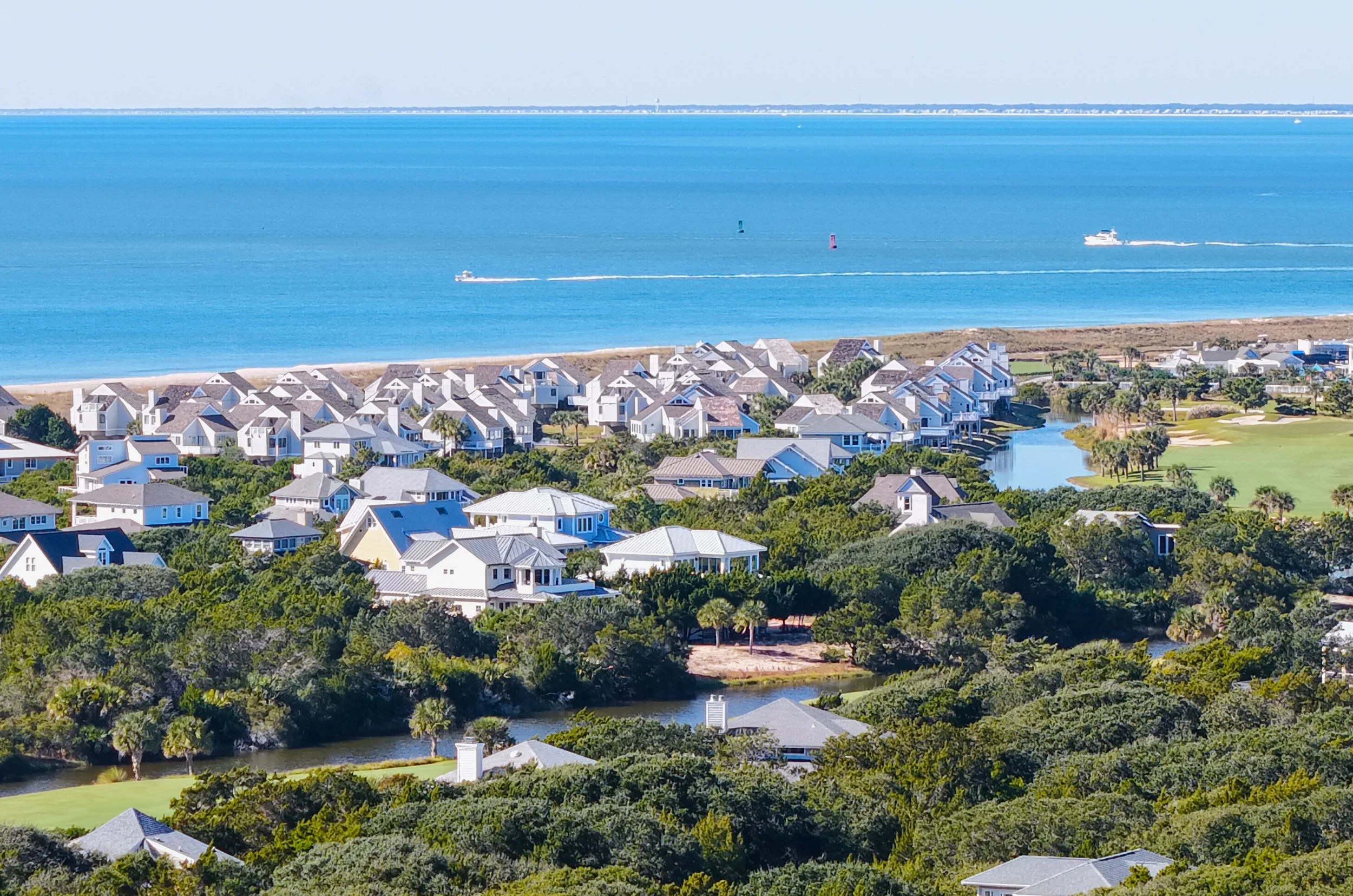 Aerial view of a coastal residential neighborhood with houses, lush green trees, a waterway, and the ocean with boats in the background.