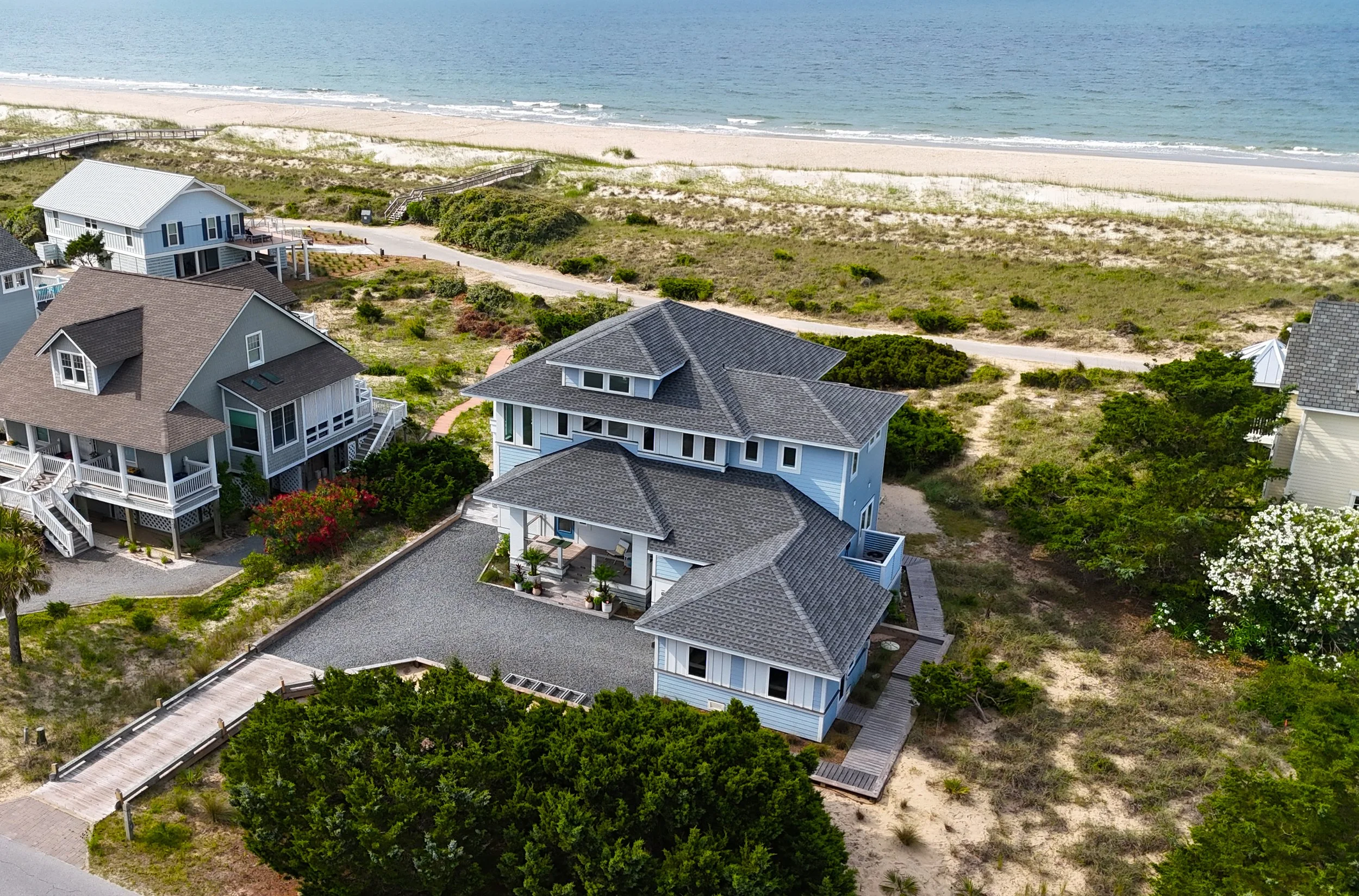 An aerial view of beach houses near the shoreline, with a sandy beach and ocean in the background, greenery around the houses, and a pathway leading to the beach.