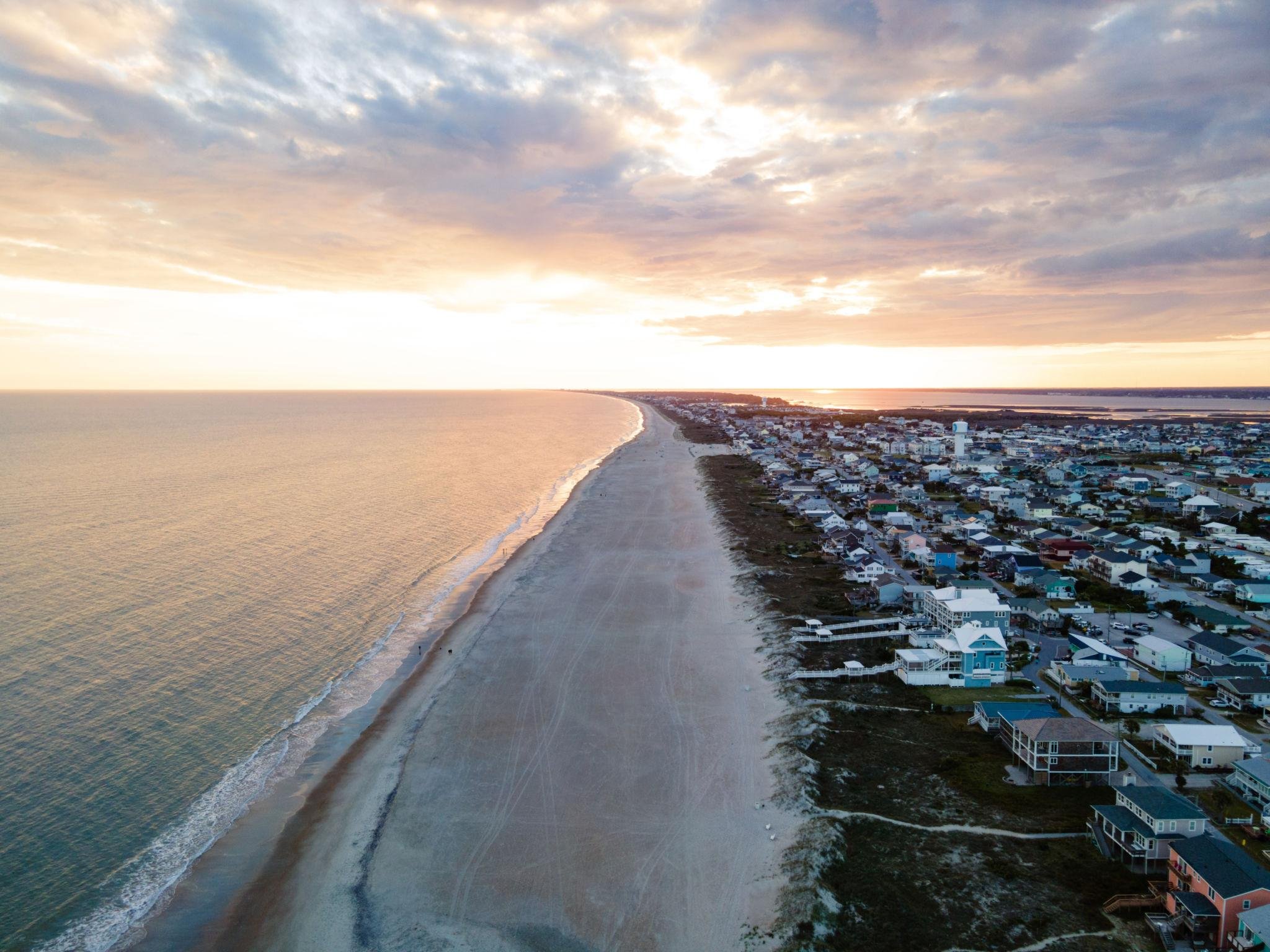 An aerial view of a beachside community at sunset, with sandy shoreline, residential houses, and calm water under a partly cloudy sky.