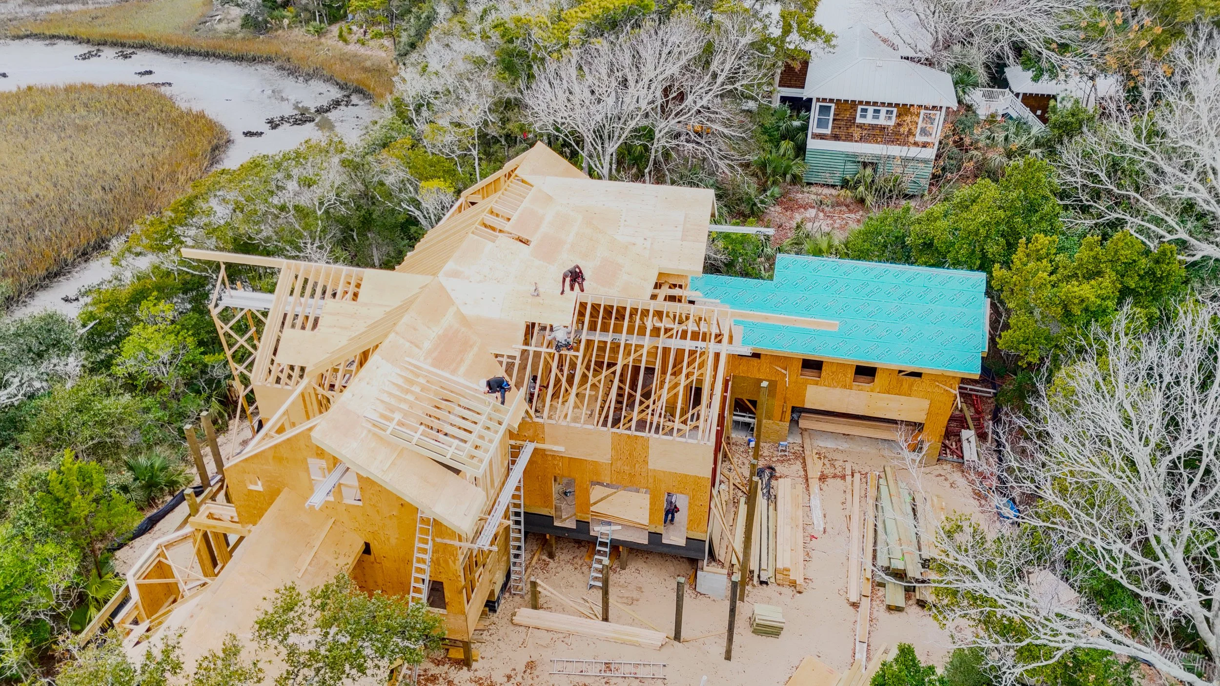 Aerial view of a house under construction surrounded by trees, with workers on the roof and a blue steel roof panel installed on part of the roof.