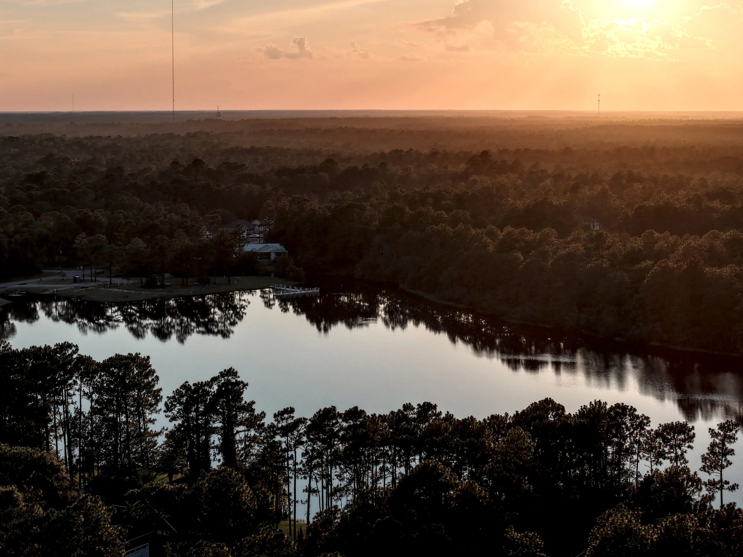 Sunset over a forested area with a body of water reflecting trees, houses, and the sky with scattered clouds.