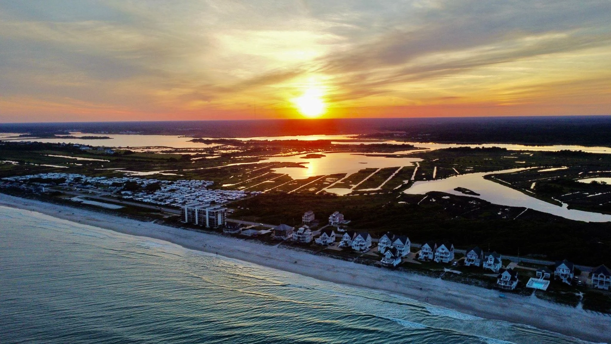 An aerial view of a coastline at sunset, featuring a beach, ocean, residential houses, and marshlands with water channels.