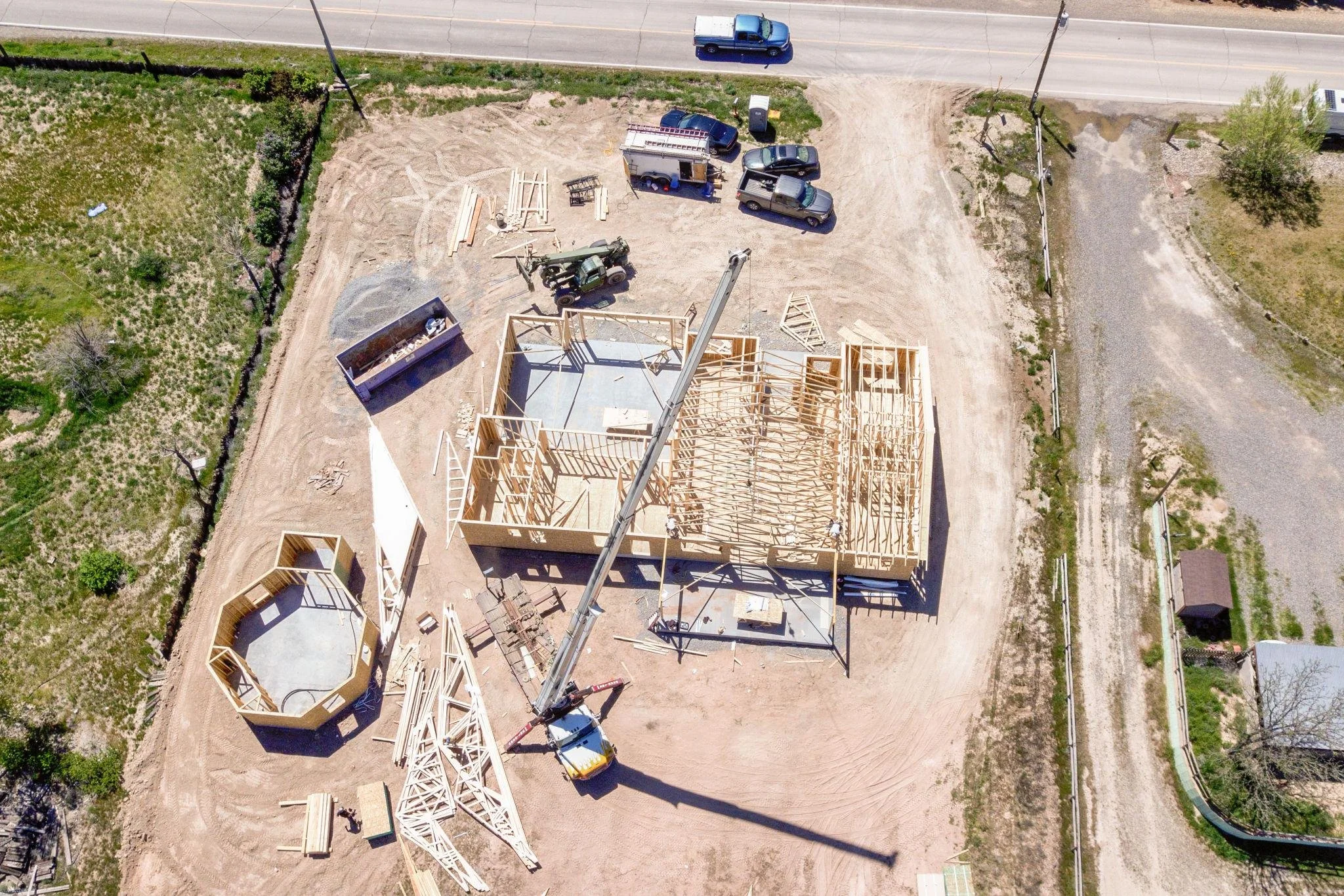 Aerial view of a construction site with wooden framing for a building, construction equipment including a crane, trucks, and piles of wood, next to a road and green areas.