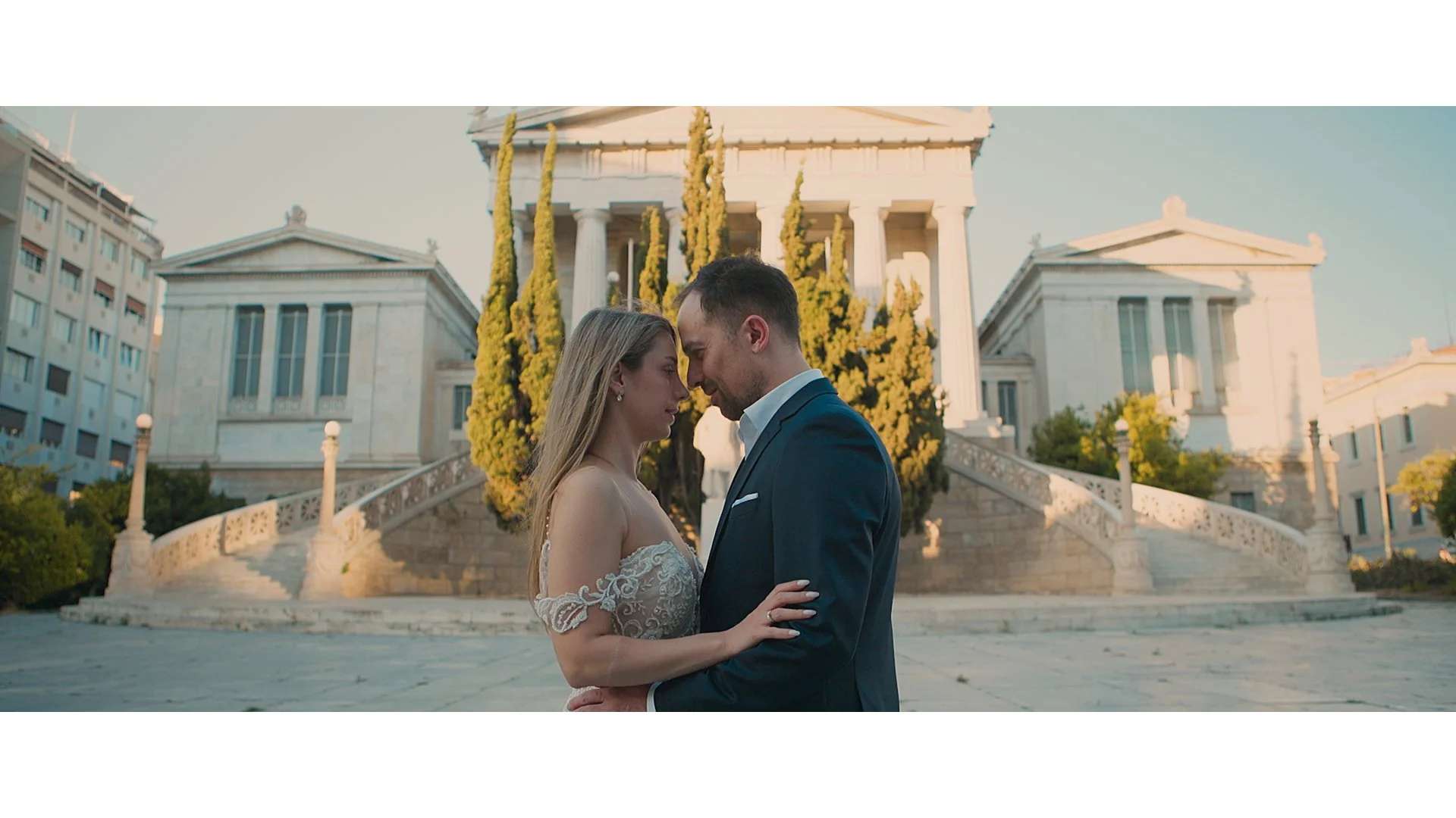 A couple on their wedding day, standing close together, with their foreheads touching, in front of a grand, classical-style building with stairs and columns, during sunset.