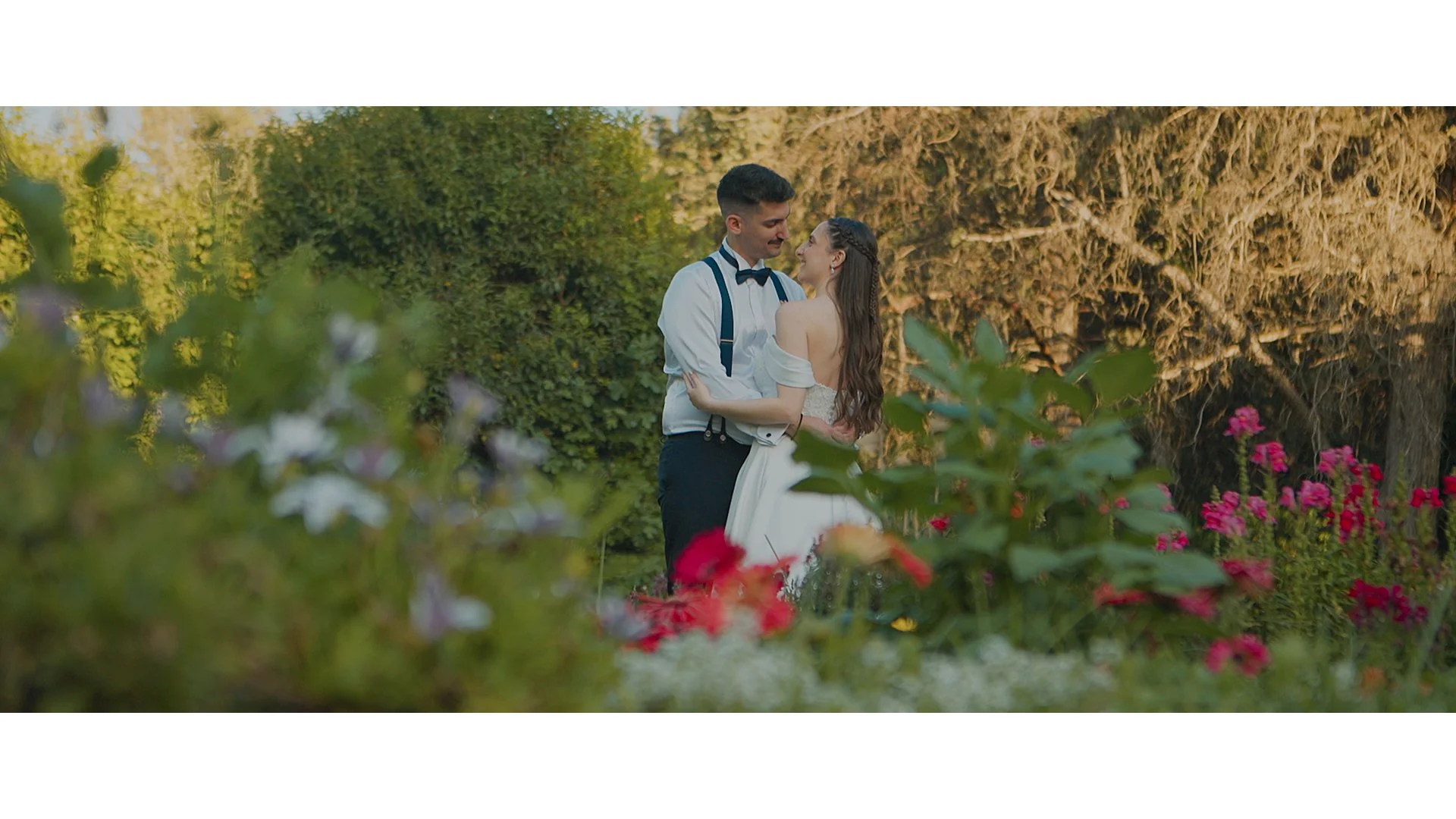 A man in a tuxedo with suspenders and a woman in a white dress dancing in a garden with lush greenery and colorful flowers.