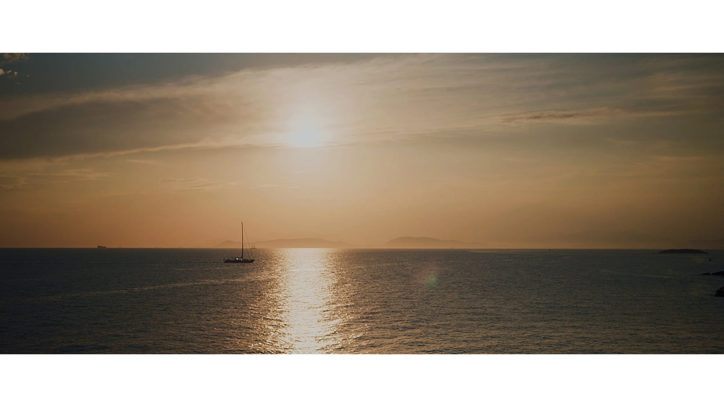 Sunset over calm ocean with a sailboat and distant islands.