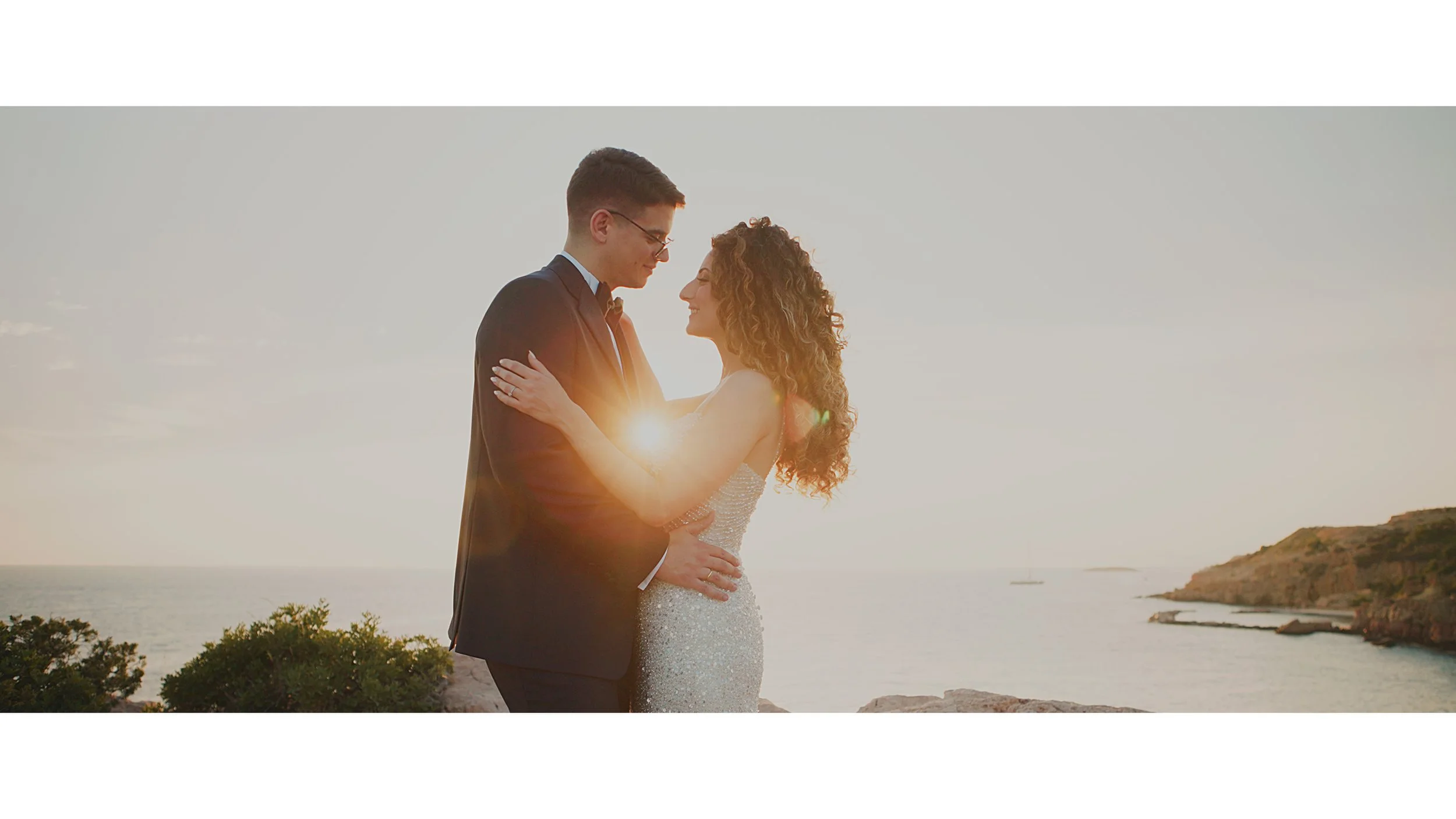 A newlywed couple facing each other and smiling at sunset near the ocean, with the woman wearing a white wedding dress and the man in a dark suit.