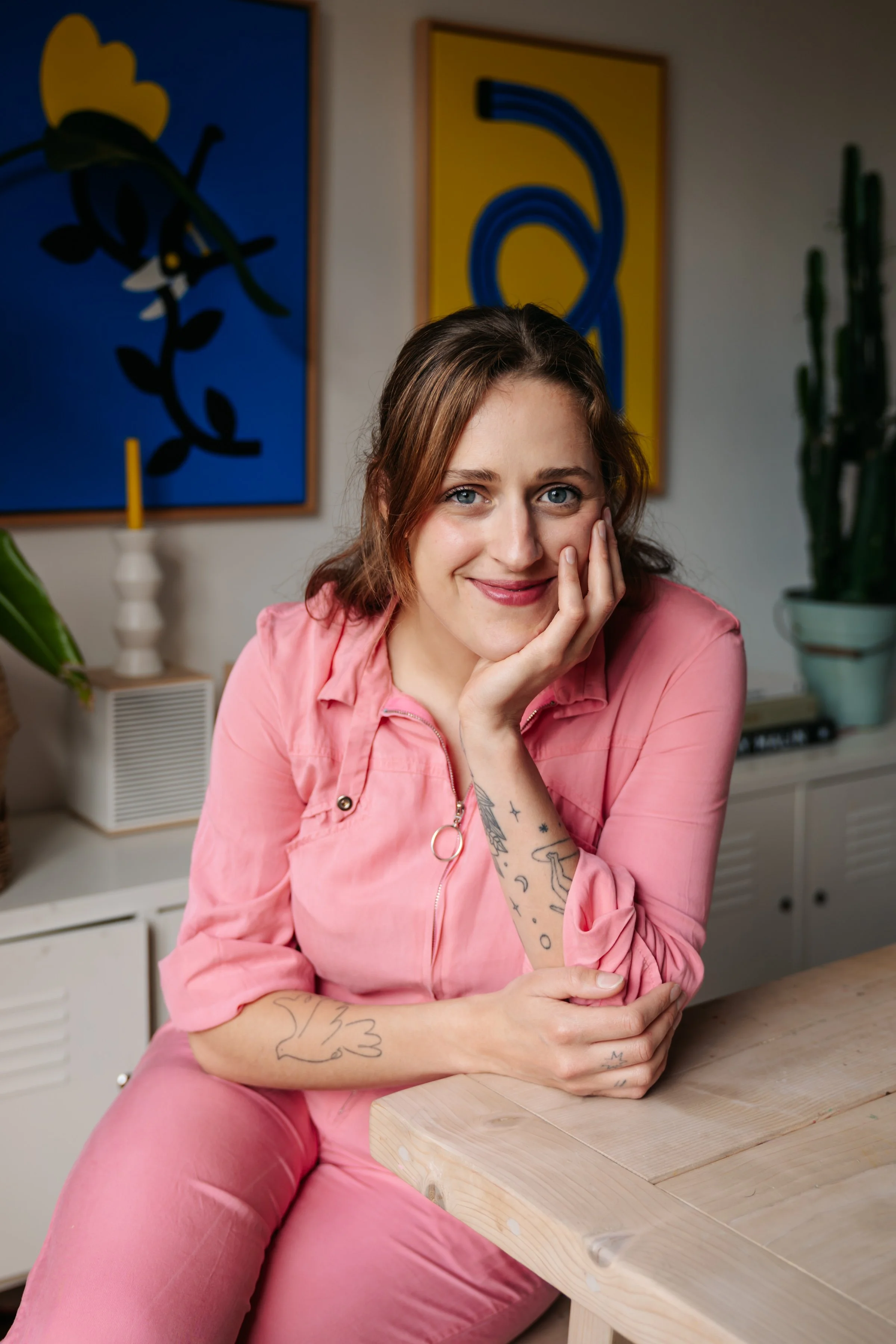 A woman with brown hair and tattoos on her arms, wearing a pink outfit, sitting at a wooden table with her head resting on her hand, smiling at the camera. Behind her are abstract art pieces and a potted cactus plant.
