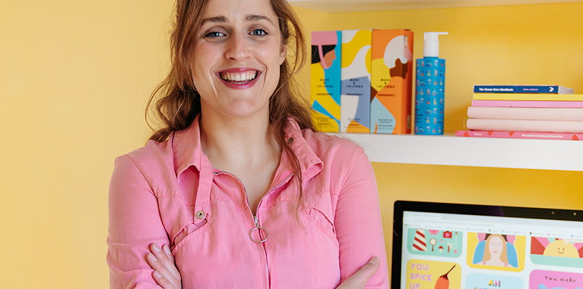 A smiling woman with brown hair wearing a pink shirt, arms crossed, standing in front of a yellow wall with a white shelf holding colorful books, a blue bottle, and a computer monitor displaying colorful graphics.