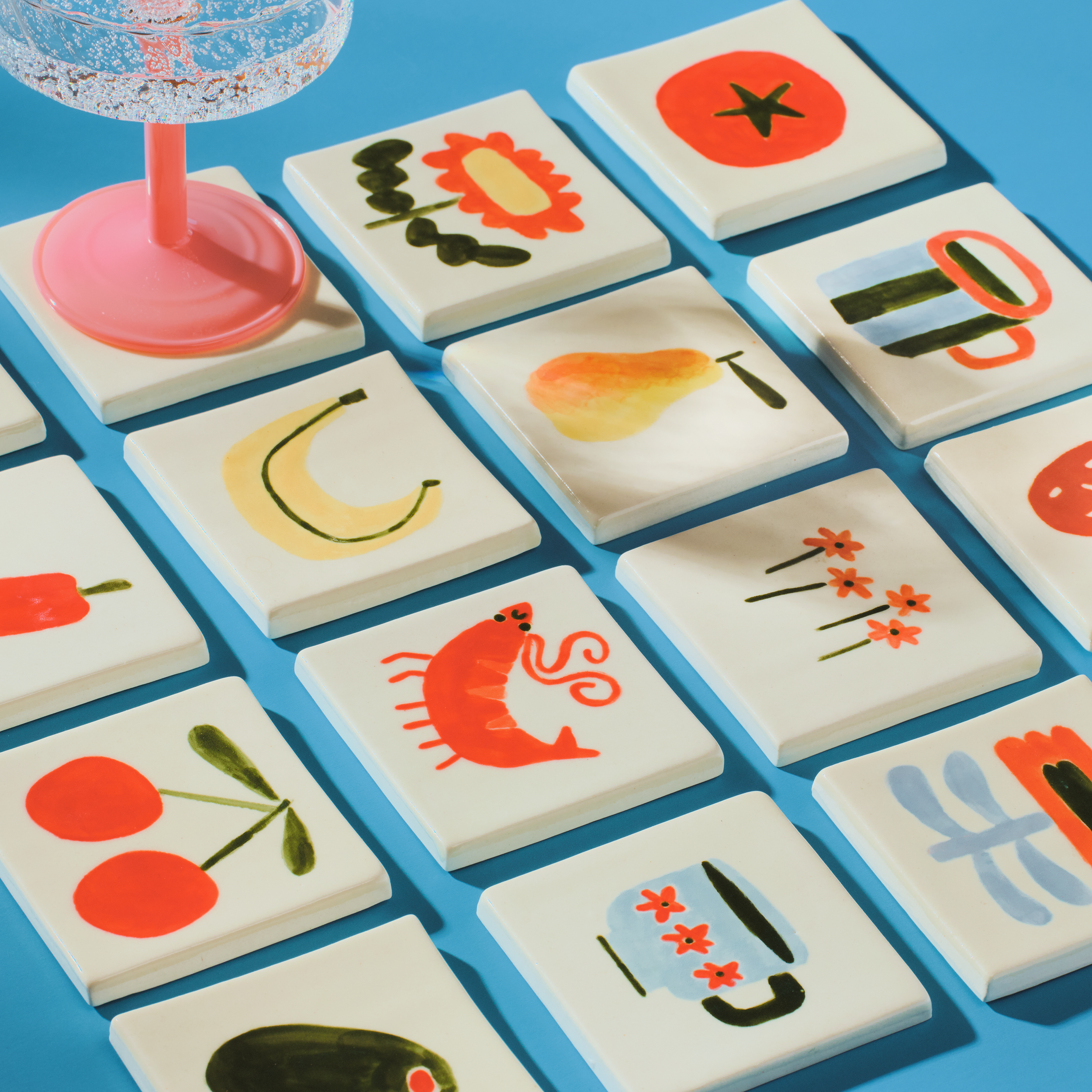 Colorful tiles with fruit and floral illustrations, placed on a blue surface, with a glass of clear bubbly water on the left.