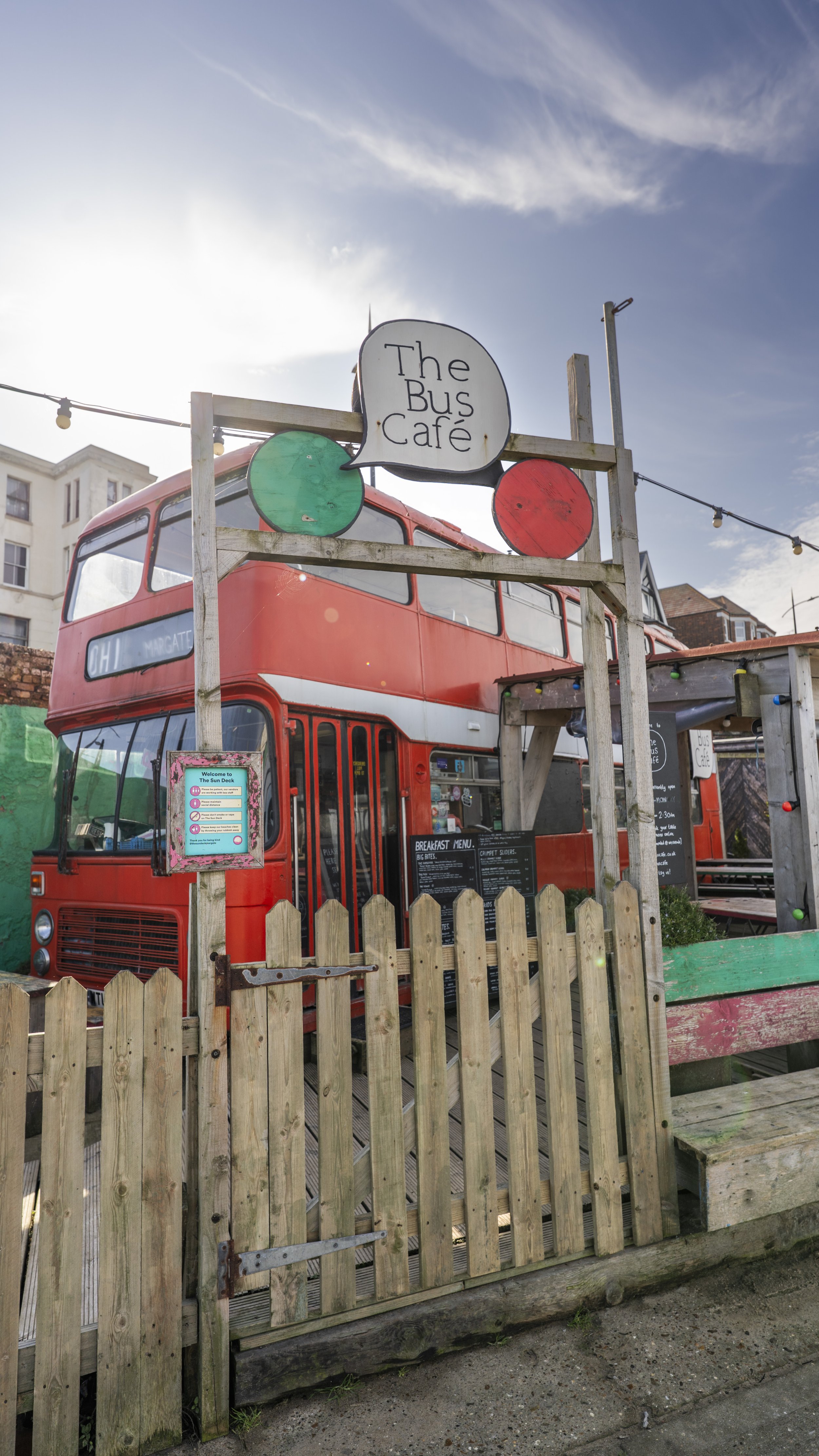 A red double-decker bus turned cafe with a sign reading 'The Bus Cafe' on a wooden frame, surrounded by outdoor seating and string lights, under a partly cloudy sky.
