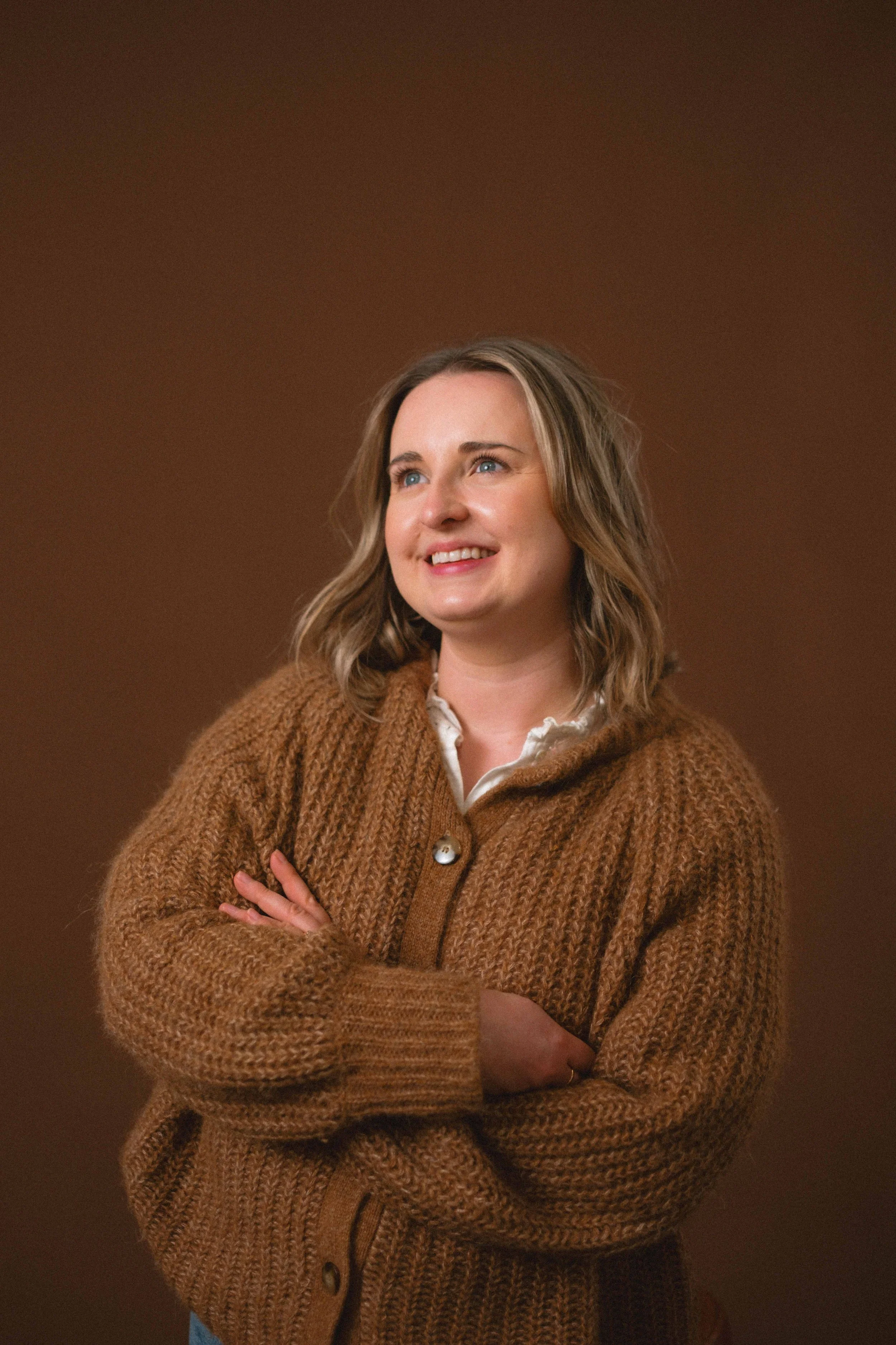 A woman with blue eyes and curly, shoulder-length hair wearing a brown knitted cardigan, standing with her arms crossed and smiling slightly against a plain brown background.