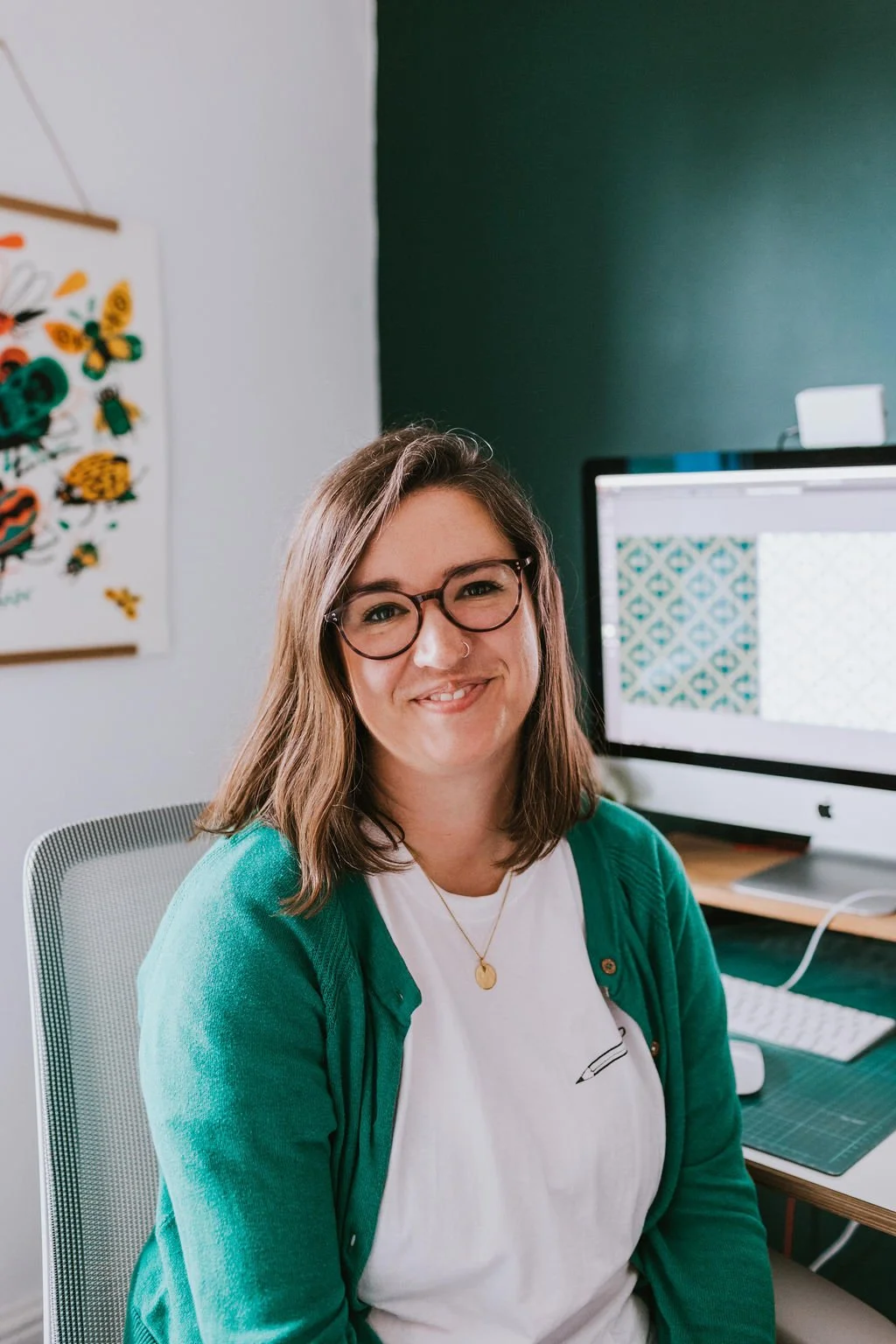 A woman with glasses and shoulder-length brown hair, smiling, sitting in an office. She is wearing a white t-shirt and a green cardigan, with a gold necklace. Behind her, there is a computer monitor and colorful artwork on the wall.