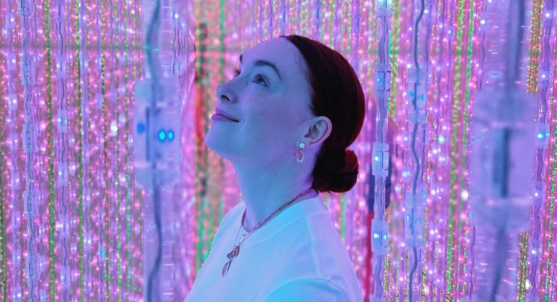 A woman with dark red hair tied back, wearing a white shirt, earrings, and necklace, standing among colorful LED light strings hanging vertically, creating a vibrant, glowing atmosphere.