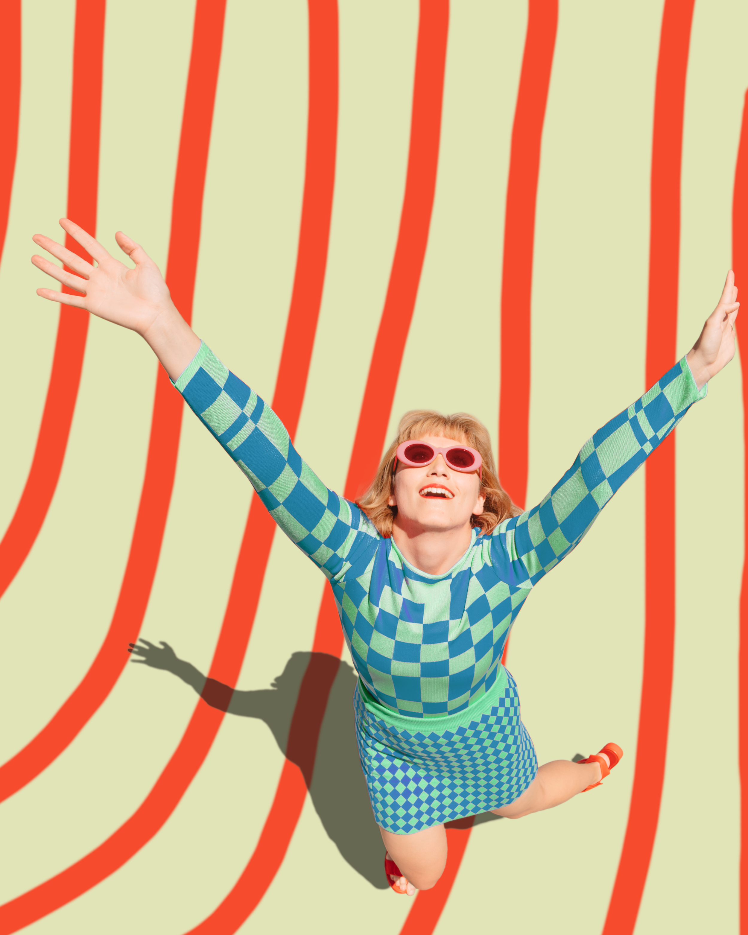 A woman with curly red hair, wearing pink sunglasses, a blue and green checkered dress, and orange shoes, jumps with her arms raised on a yellow background with red wavy lines.