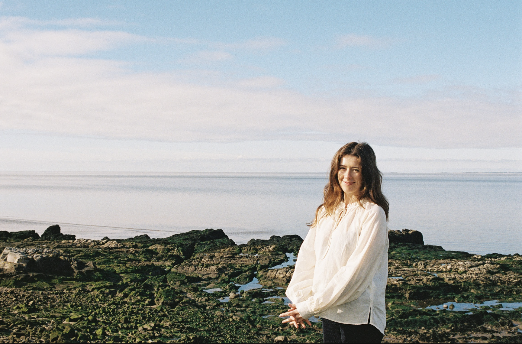 A woman in a white blouse standing on a rocky shore near the water, smiling at the camera with a partly cloudy sky over the ocean in the background.