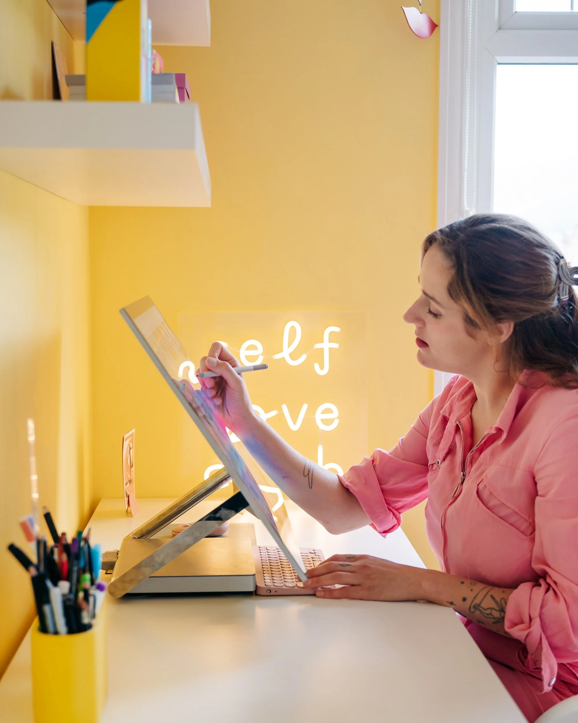 A woman with tattoos on her arm, wearing a pink shirt, sitting at a yellow desk, stylishly bangs her head in front of a computer. The background behind her is yellow, with a window on the right and decorative items on the shelf.