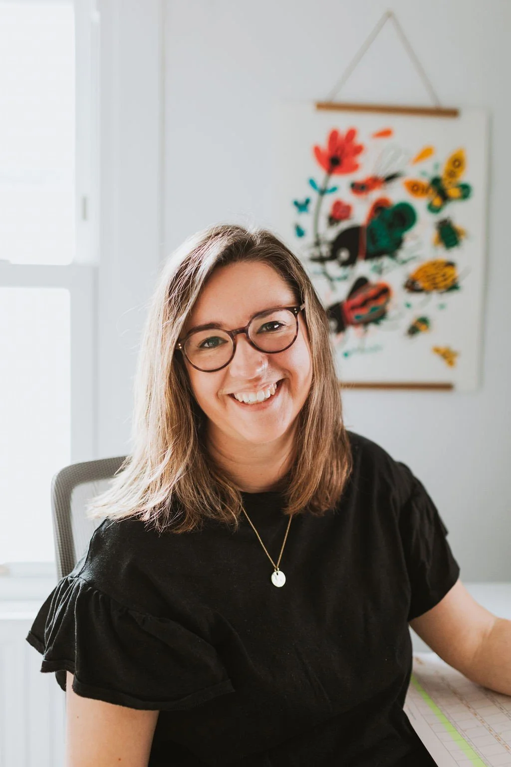A woman with shoulder-length brown hair, glasses, and a black shirt smiling at the camera indoors.