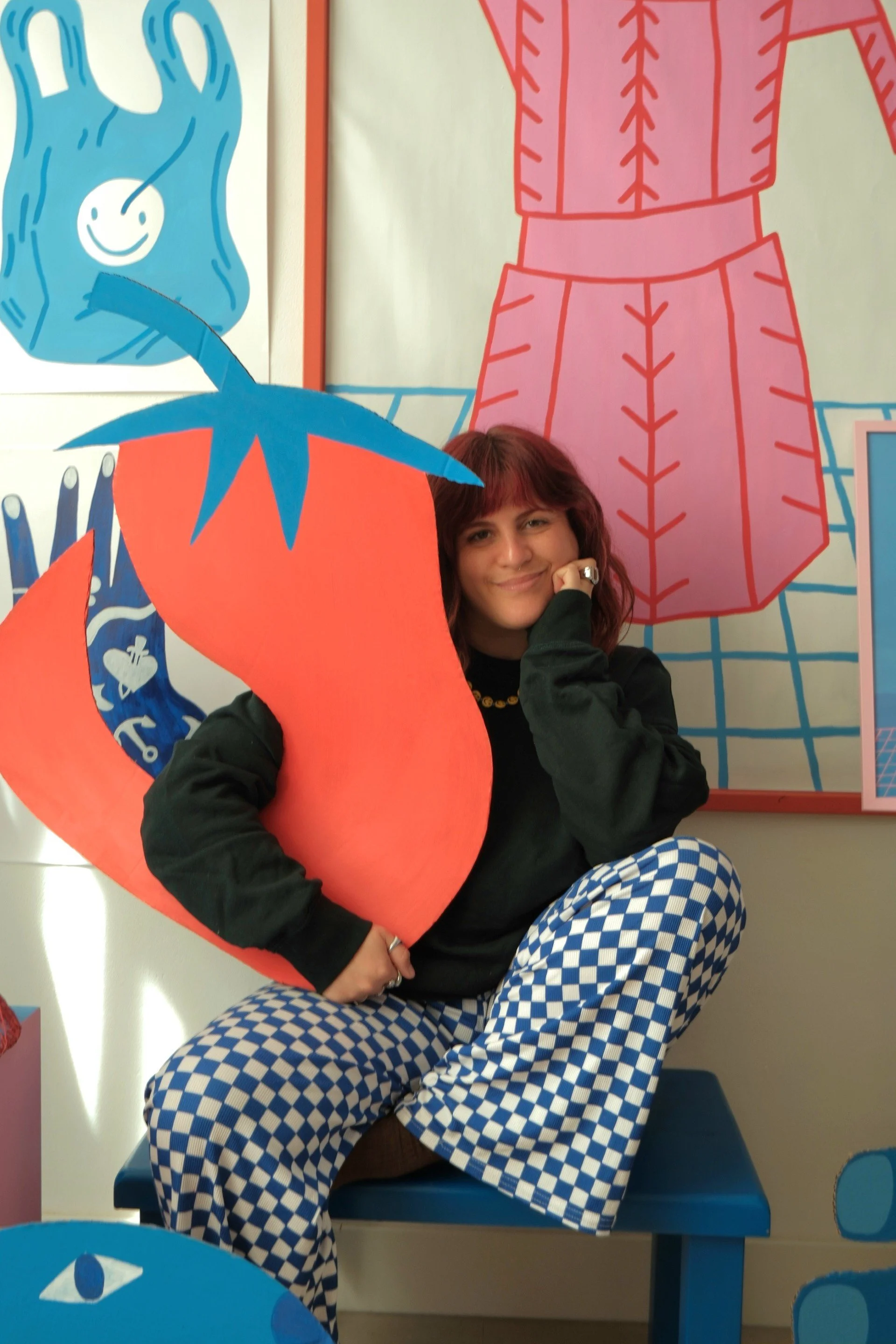A woman with red hair, smiling, sitting on a blue chair in front of a colorful, cartoonish mural. She is wearing a black hoodie and blue-and-white checkered pants, holding a large orange and blue paper flower.