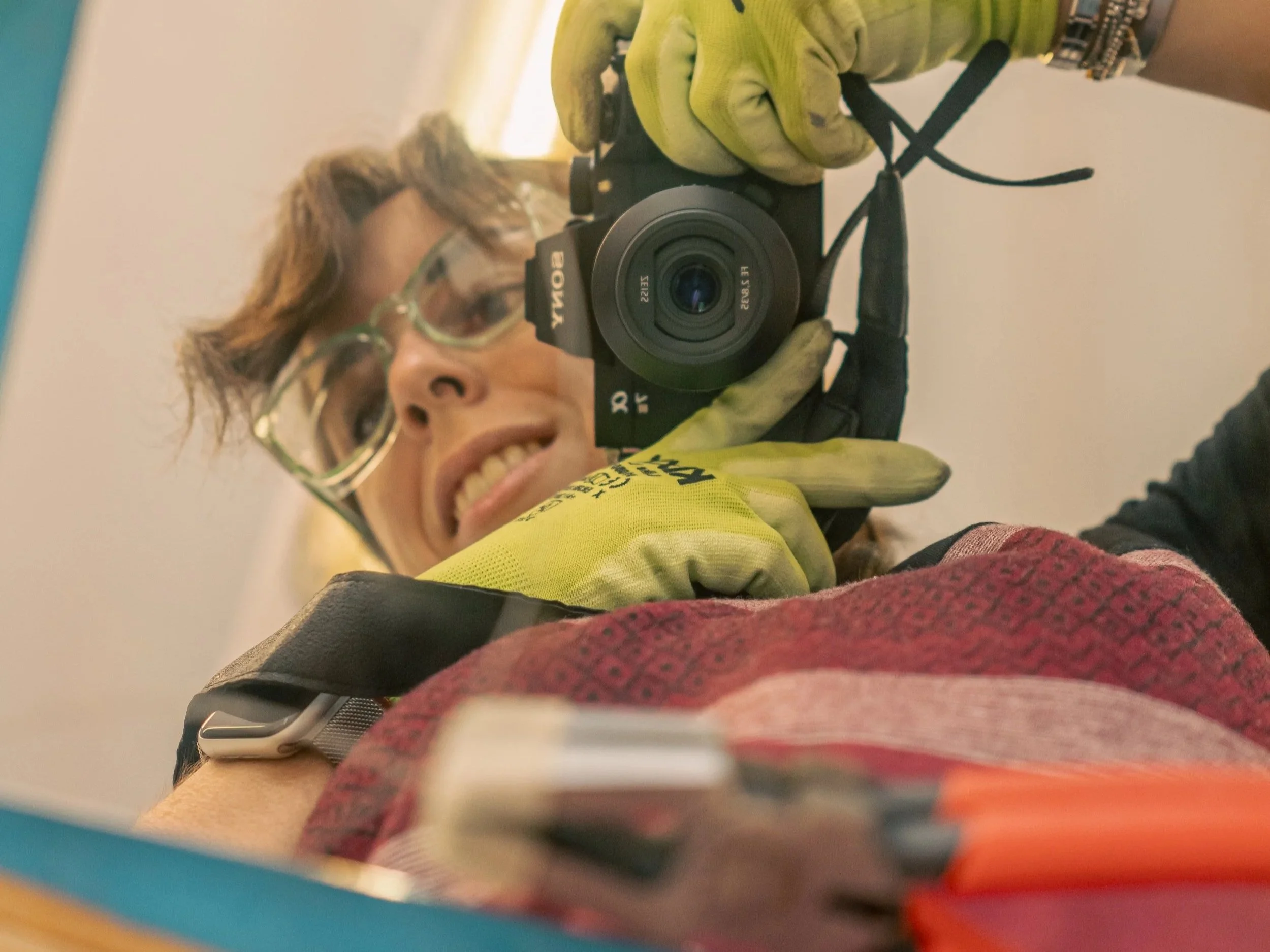 A person with light brown hair, glasses, and a yellow jacket is taking a photo with a camera, reflected in a mirror. Glass working tools are also reflected.
