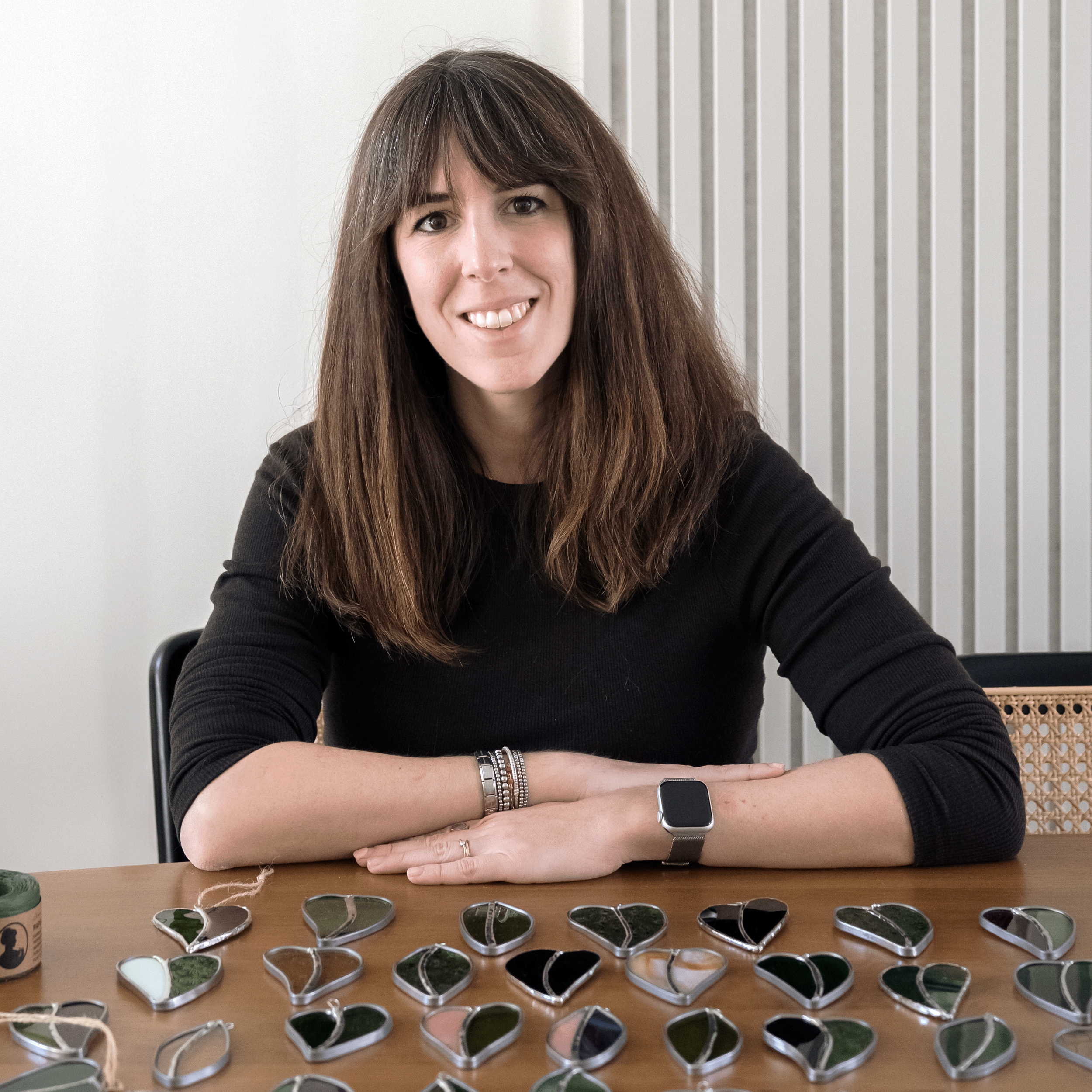 Woman with long brown hair and a watch smiling at the camera while sitting at a table with various small stained glass ornaments.