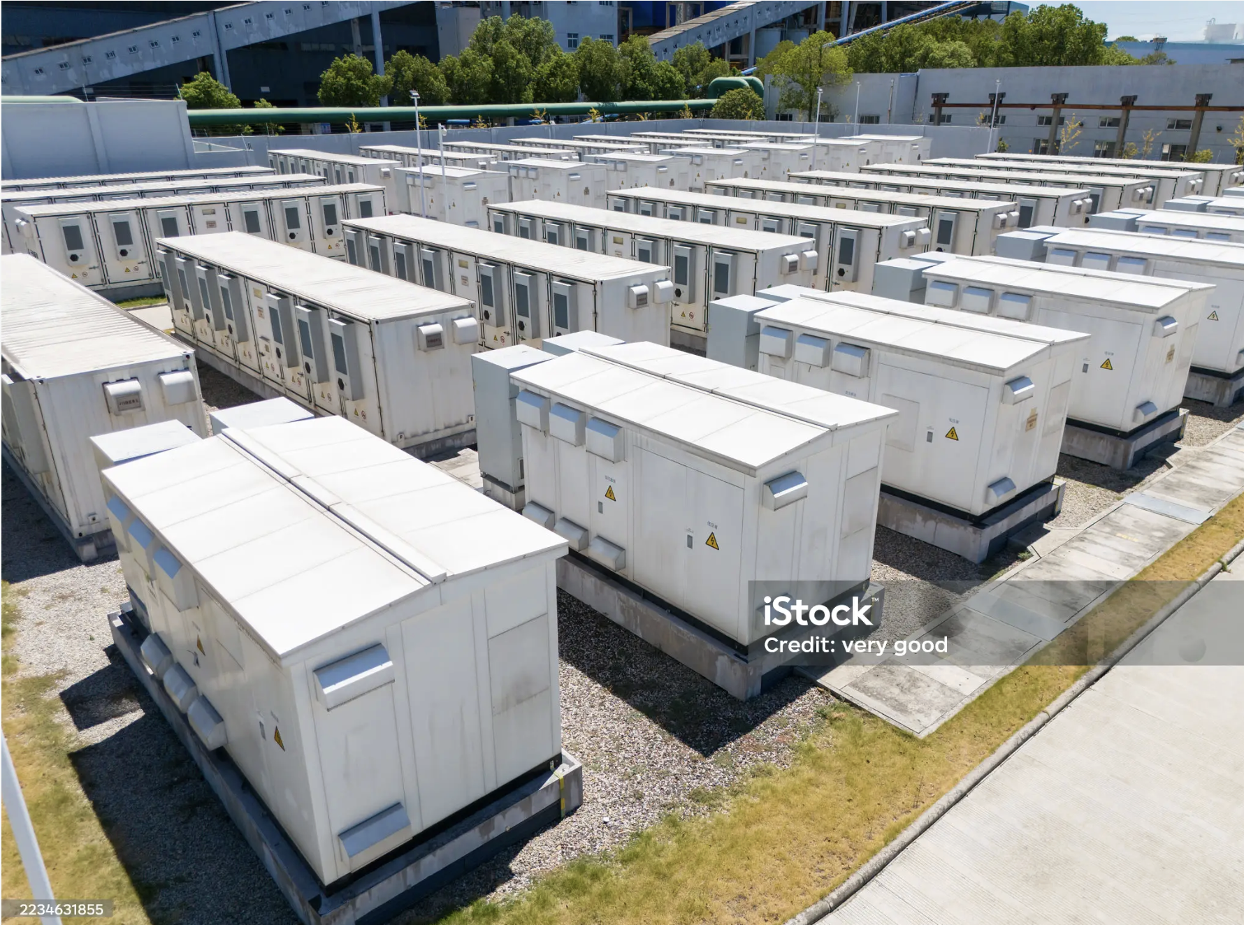 A large outdoor area filled with rows of white electrical or HVAC units with warning labels, in front of a modern building and green trees.