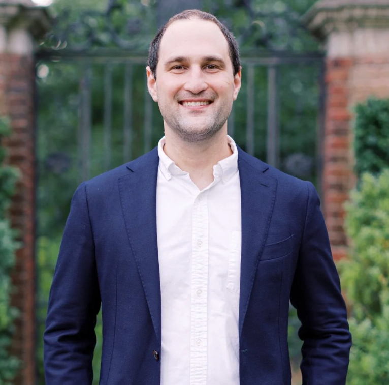 A smiling man standing outdoors in front of a garden with greenery and brick structures, wearing a white shirt and navy blue blazer.