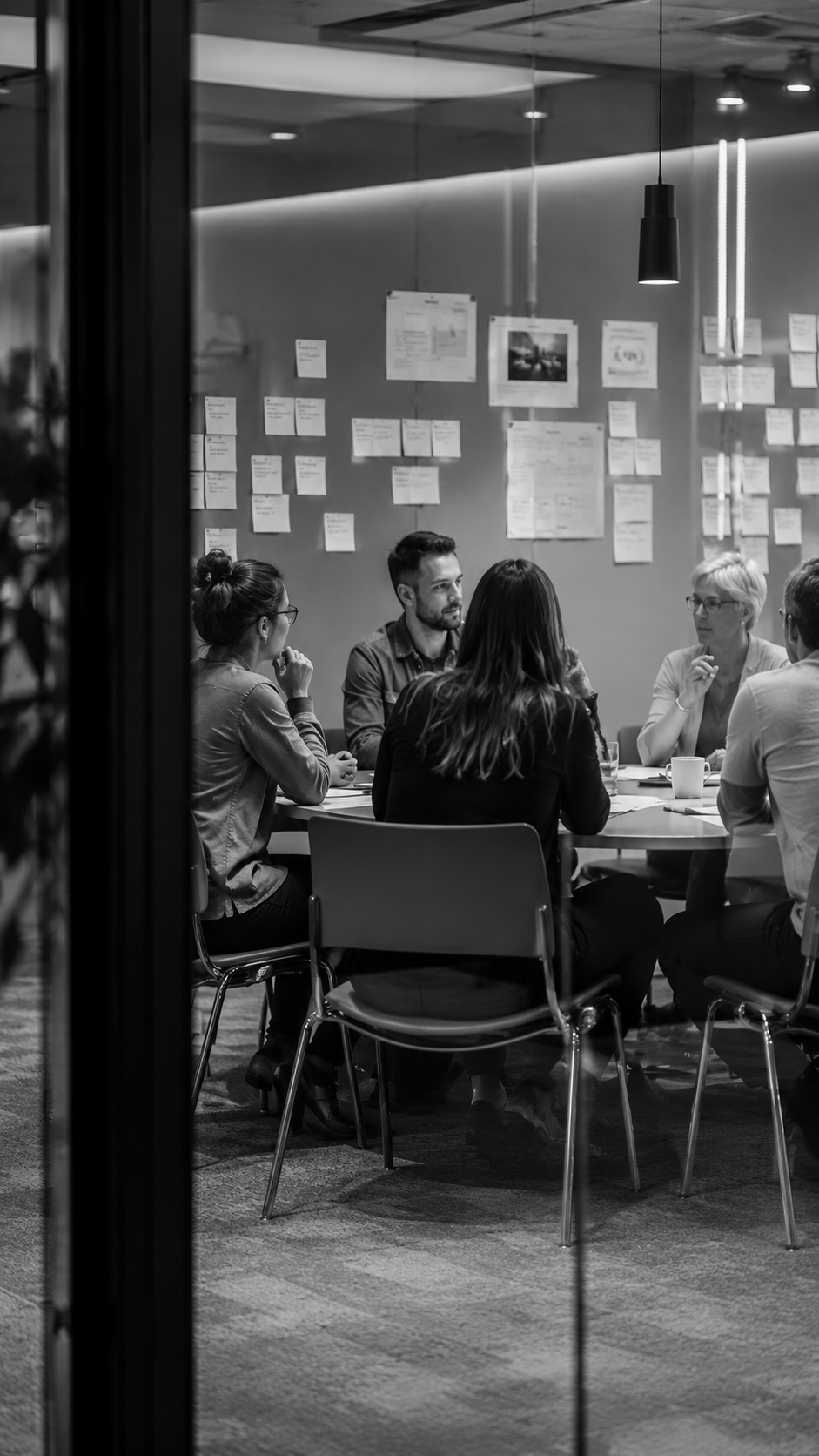 A black-and-white photo of five people sitting around a table in a modern conference room, engaged in discussion, with notes and papers on the wall behind them.
