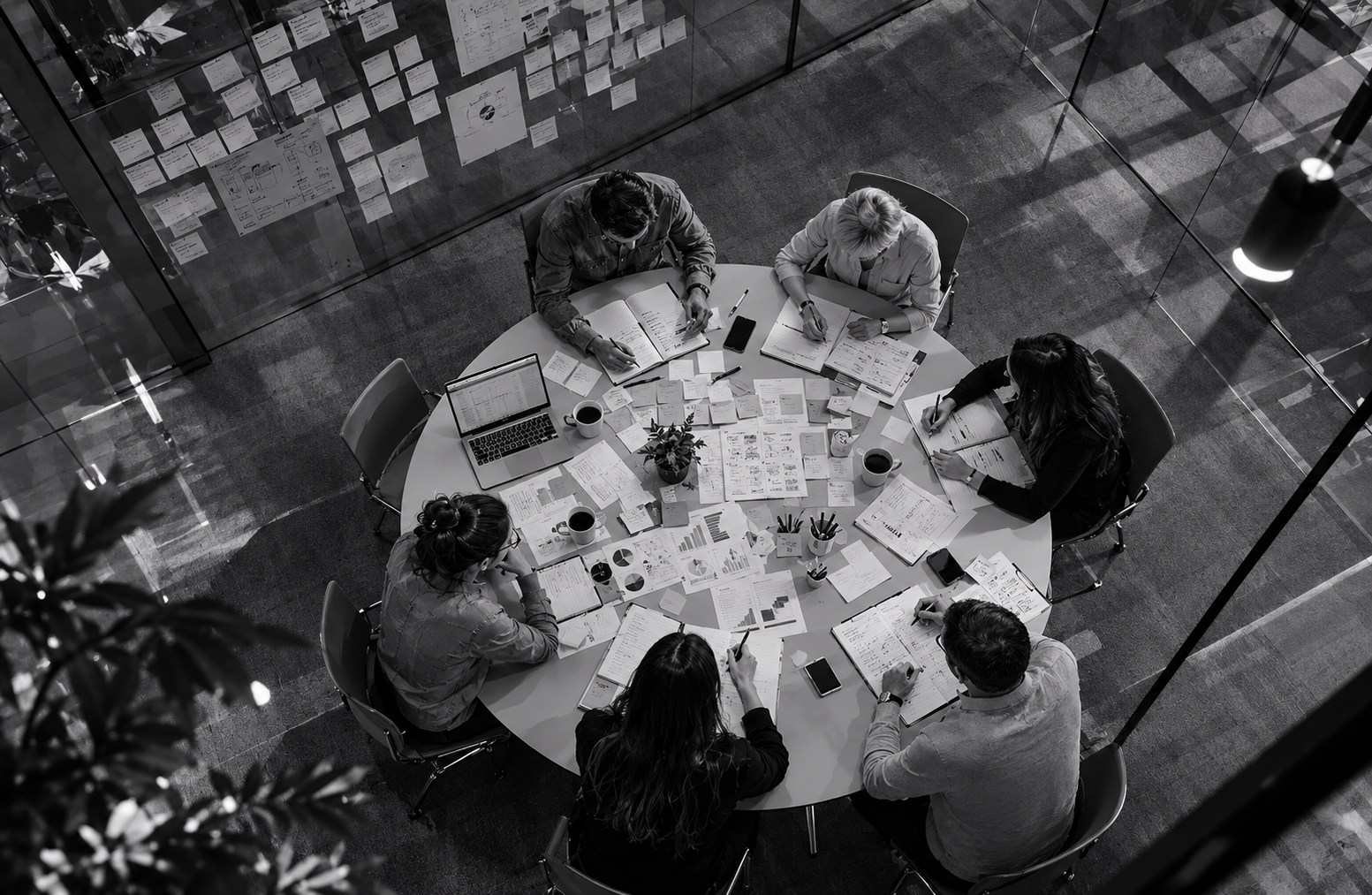 Six people sitting around a round table covered with papers, documents, graphs, laptops, and coffee cups, engaging in a meeting or discussion in an office setting.