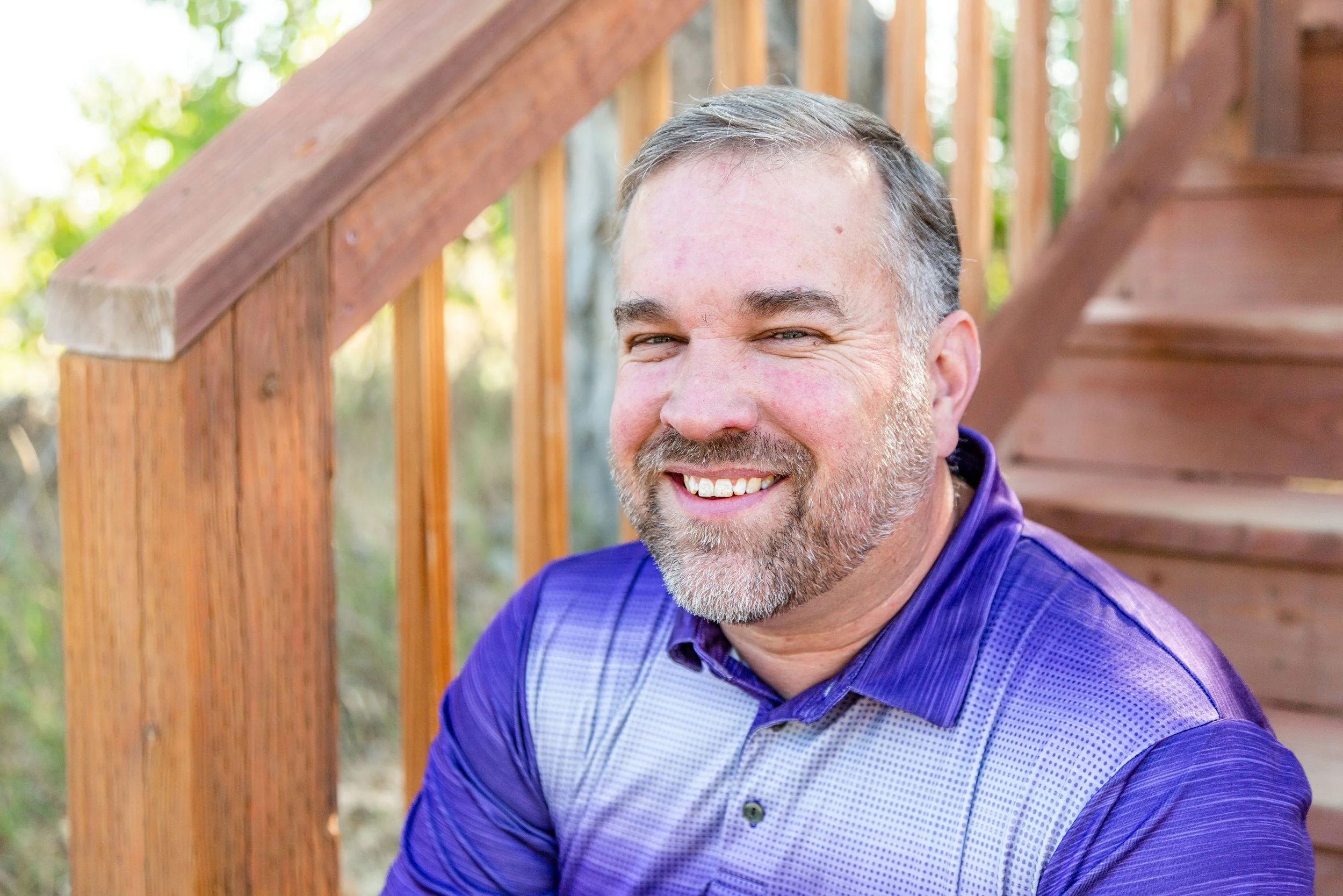Close-up of a middle-aged man with gray hair and beard smiling outdoors, leaning against a wooden fence, wearing a purple shirt.
