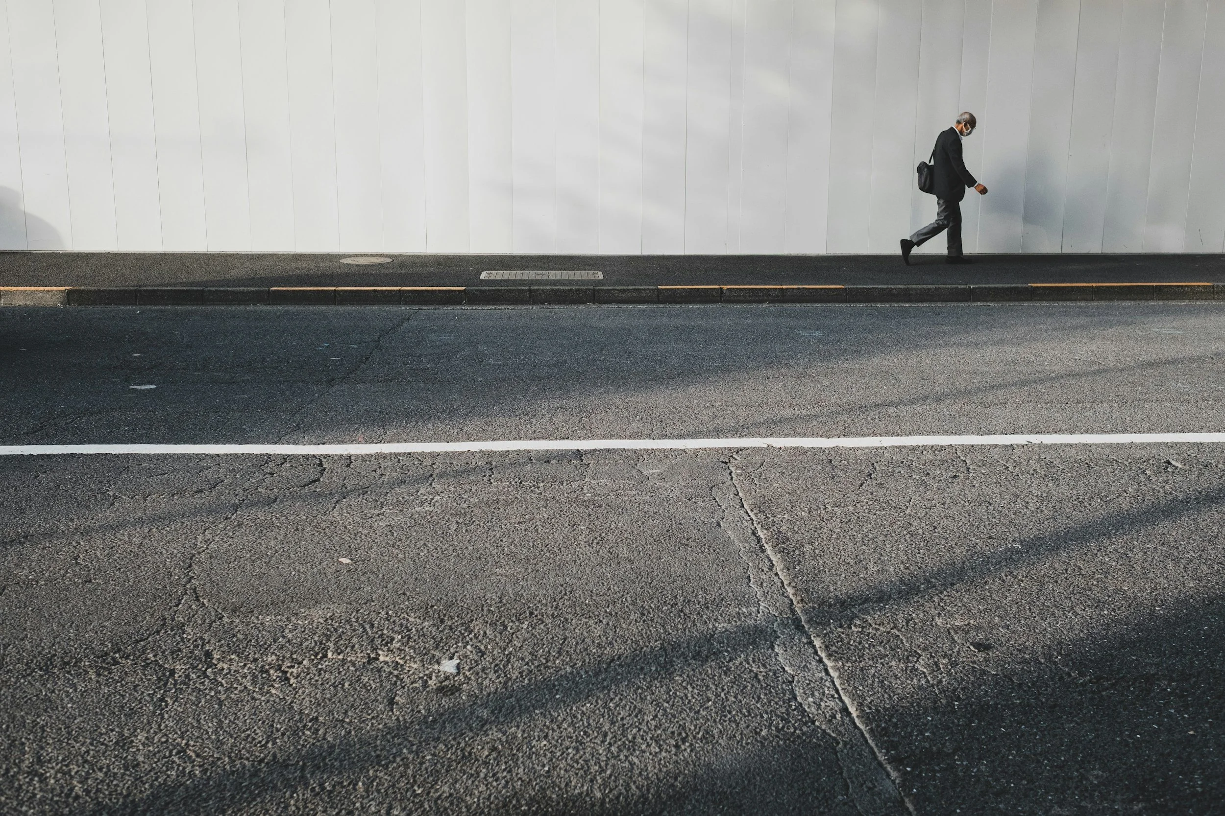 A man in a suit, wearing a face mask, walking on a sidewalk next to a plain white wall.