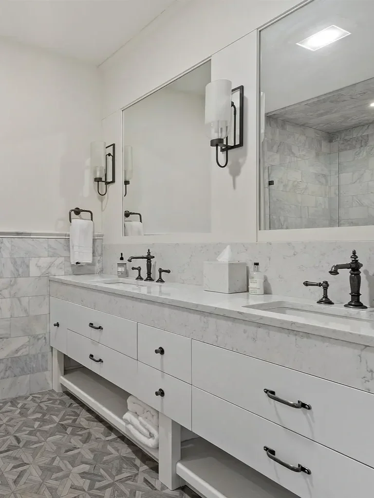 A modern bathroom vanity with a white marble countertop, black faucet fixtures, a large mirror, light fixtures, and a white cabinetry with storage drawers and an open shelf with towels.
