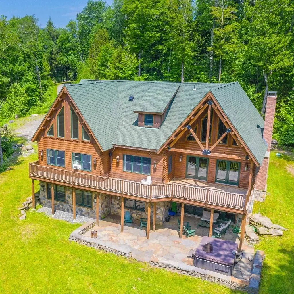A large wooden house with a green gabled roof, surrounded by tall green trees, featuring a spacious wooden deck and stone patio.