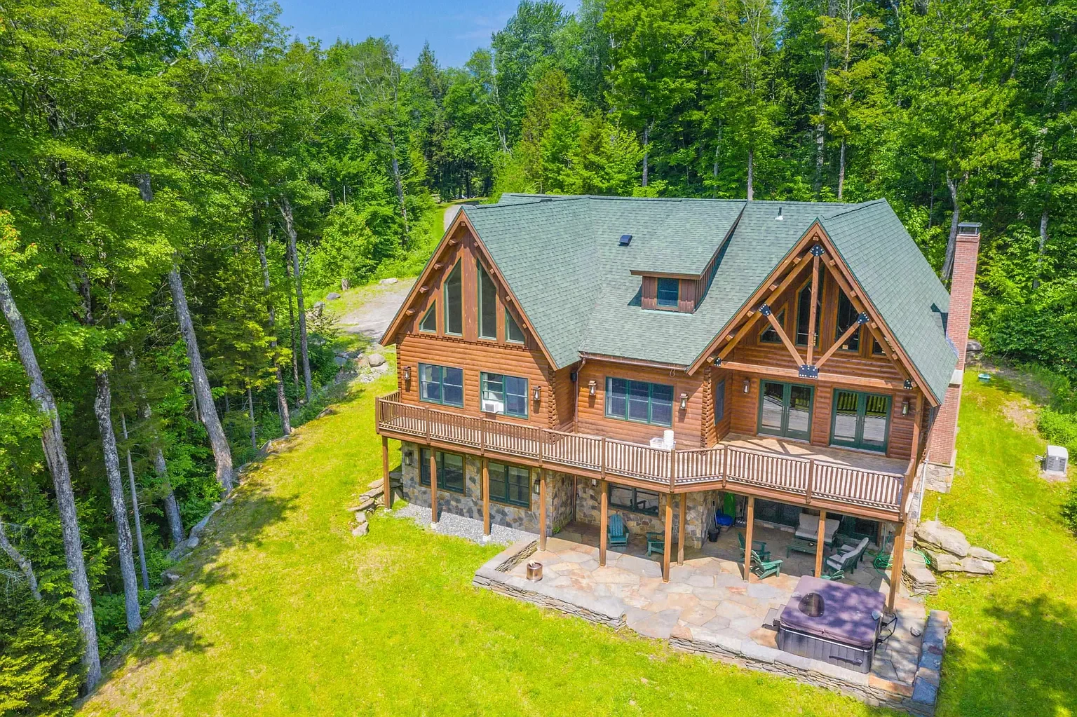 A large wooden house with a green roof surrounded by green trees, featuring a deck and a patio area.