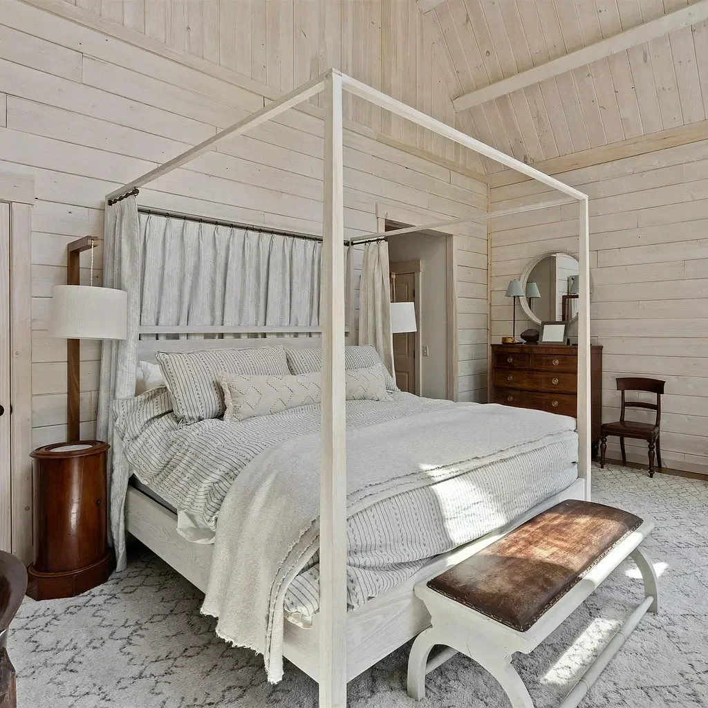 A bedroom with a white canopy bed, beige striped bedding, and light-colored wood panel walls. There is a bench at the foot of the bed, a wooden dresser with a mirror, and a small wooden chair.