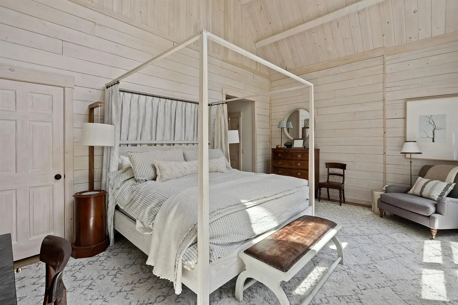 Bedroom with light-colored wooden walls and a large four-poster bed with white bedding and pillows, a small bench at the foot of the bed, a dark wooden dresser with a mirror, a gray armchair with a throw pillow, and a small side table with a lamp.