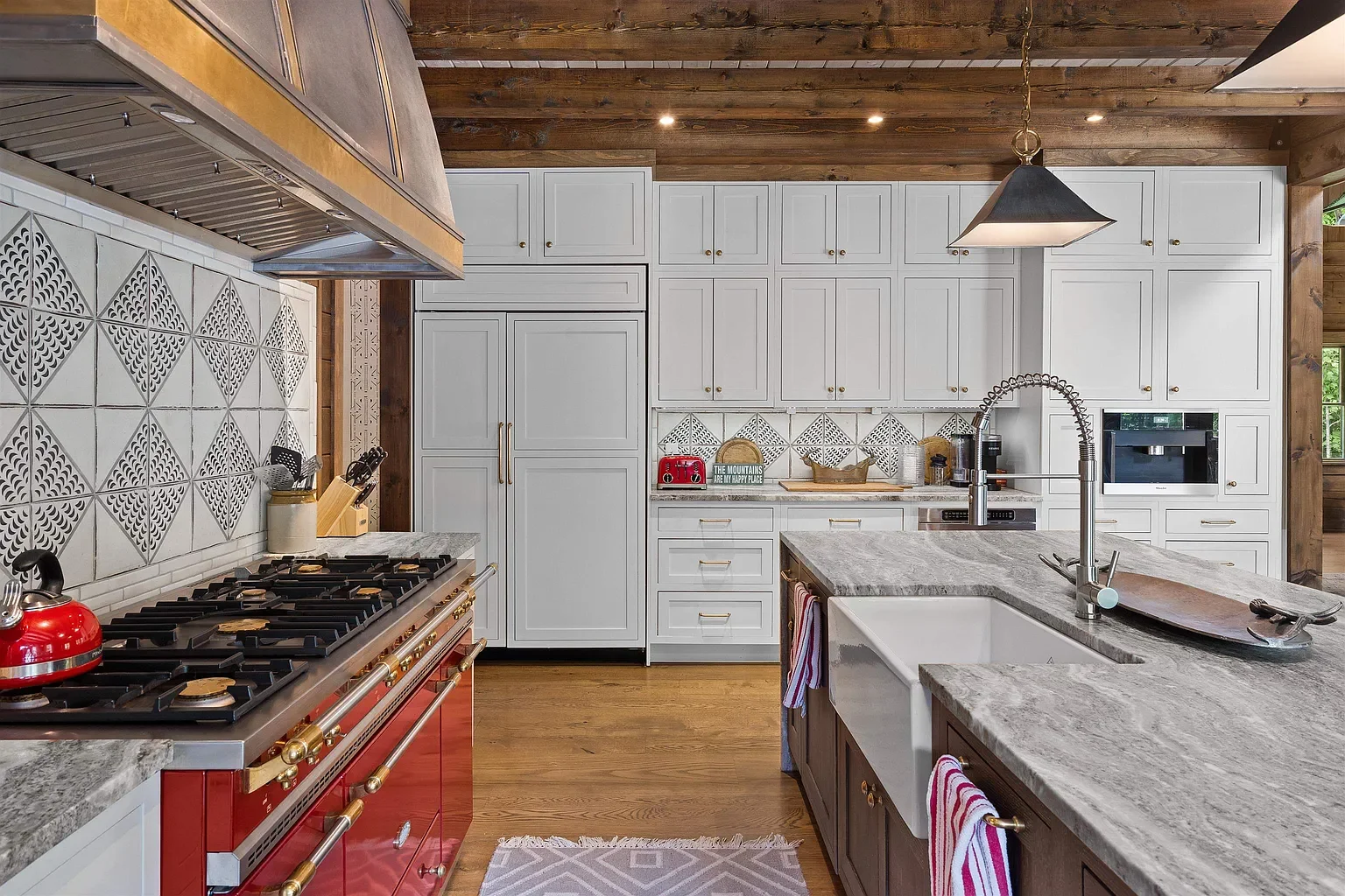 Modern kitchen with white cabinets, a gray stone countertop, a red stove, and wooden accents. There is a sink with a high-arc faucet, and a kitchen island with a gray surface. The backsplash has decorative tiles with geometric patterns. A hanging pendant light and a wooden ceiling are visible.